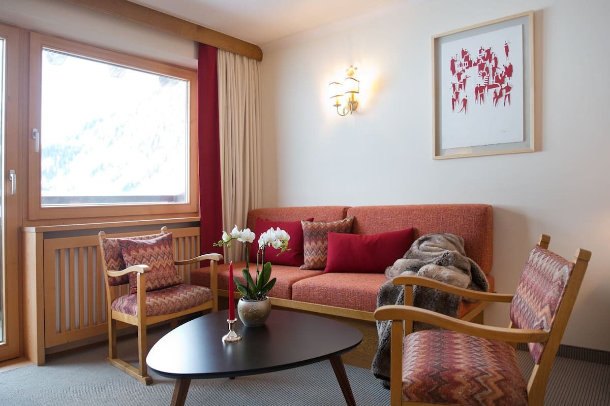 A room with a sofa, two chairs, a table with a vase and candles, a window with curtains, a wall-mounted light, and a framed picture.