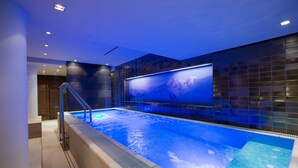 A modern indoor swimming pool with a glass wall and a large screen displaying a mountain scene. - Hotel Angela (Lech am Arlberg)