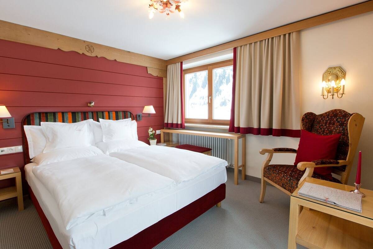 A hotel room with a large bed, a desk, a chair, a window with curtains, and a wall-mounted lamp.