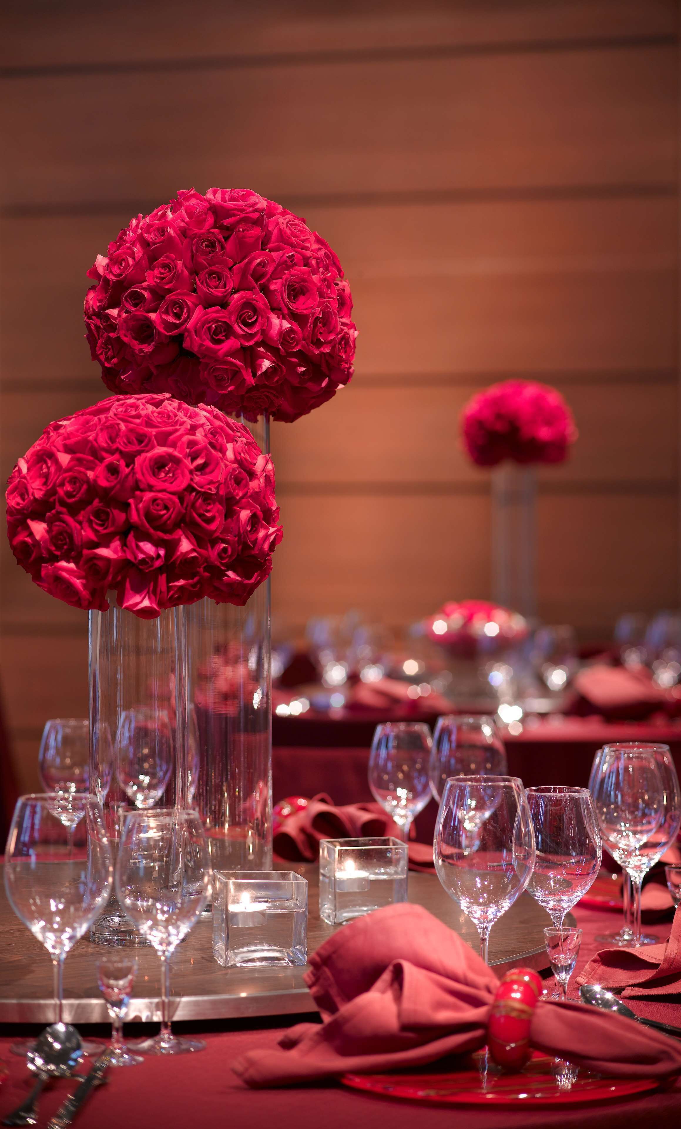 A table set with red roses in tall glass vases, clear wine glasses, and red napkins.