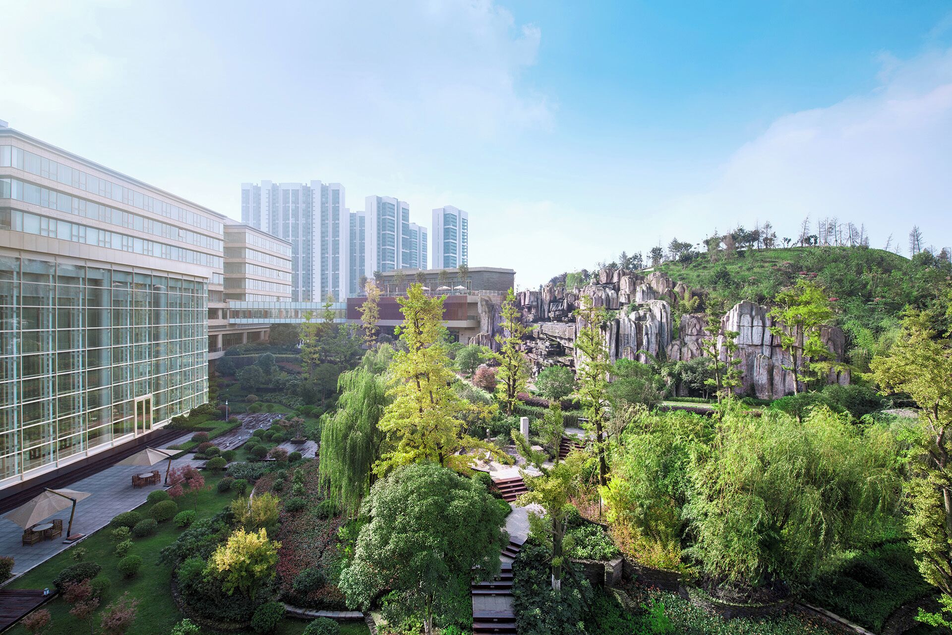 A modern building with a glass facade, surrounded by lush greenery and a rocky hill.