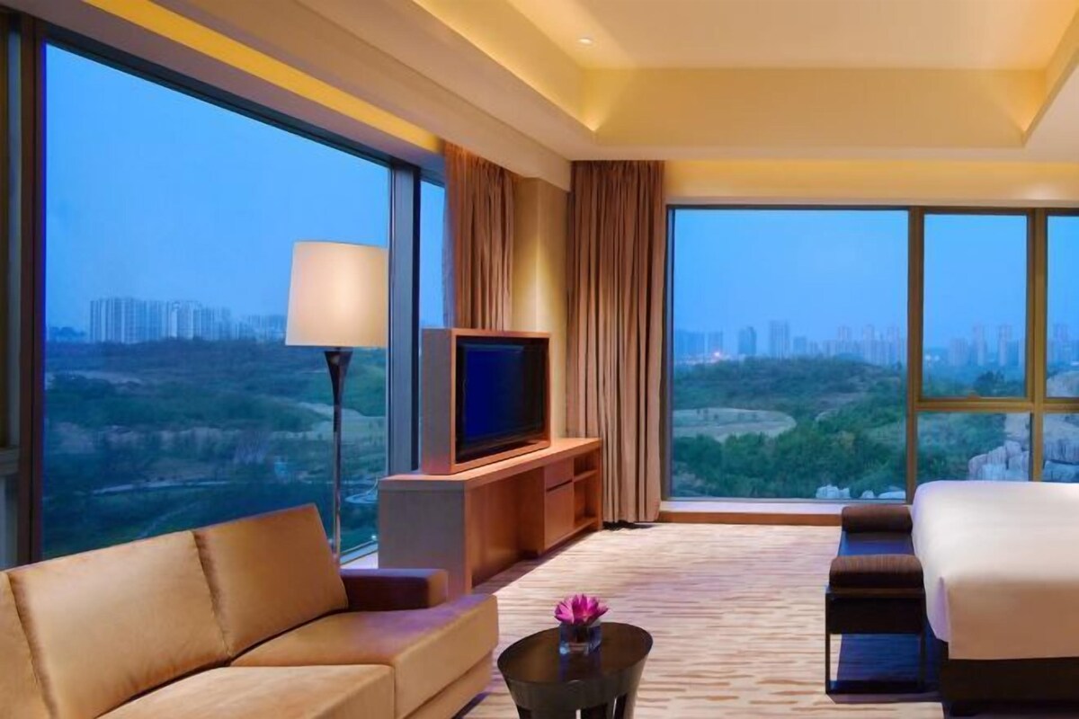 Una habitación de hotel moderna con un ventanal grande que ofrece vista a la ciudad, un televisor de pantalla plana, un sofá, una cama y una mesita de café con un florero.