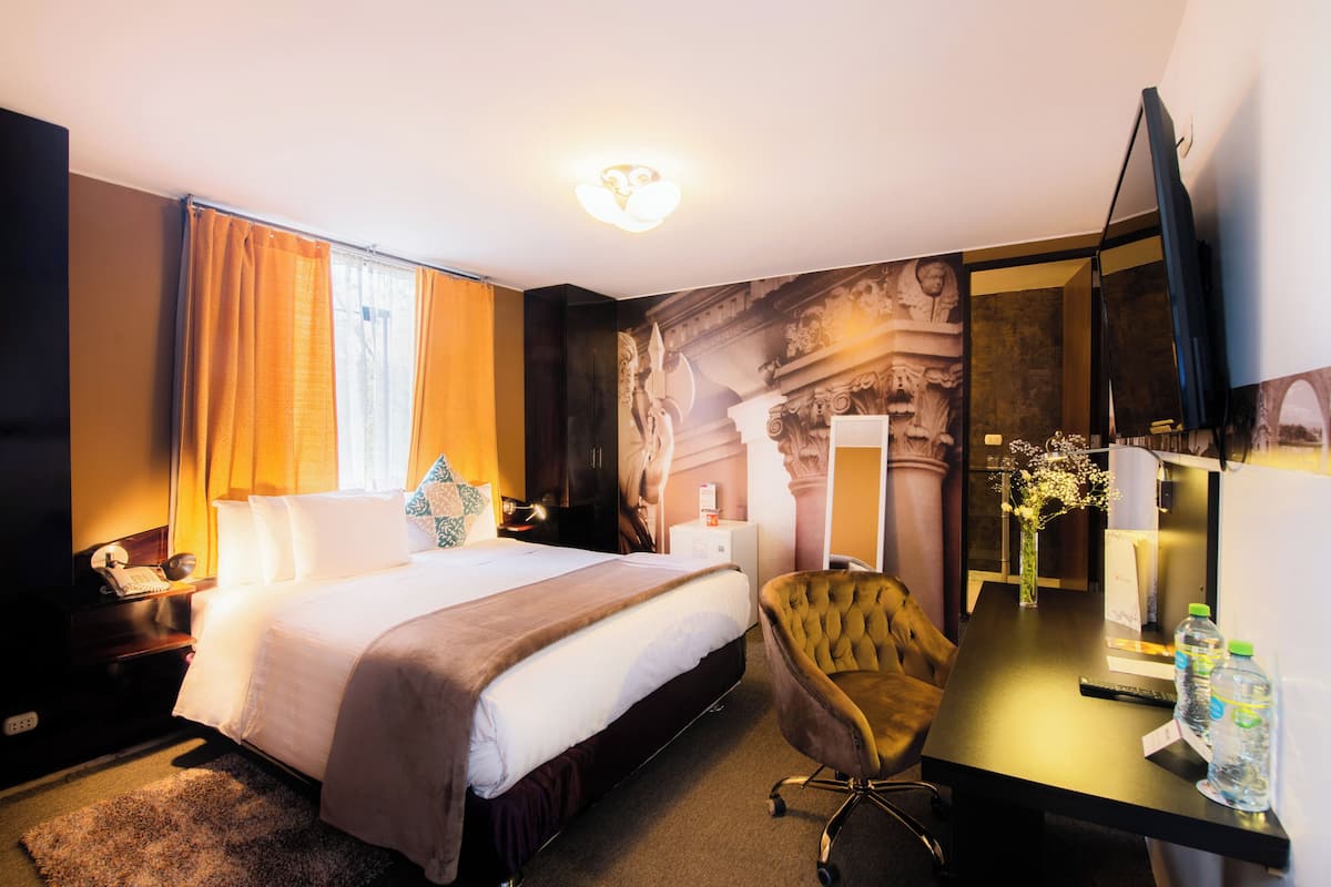 Ett hotellrum med en stor säng, ett skrivbord med en spegel, en stol och ett litet bord med en vas med blommor.