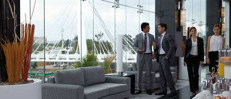 A modern hotel lobby with a grey sofa, a large window with a view of a bridge, and a buffet area with pastries.