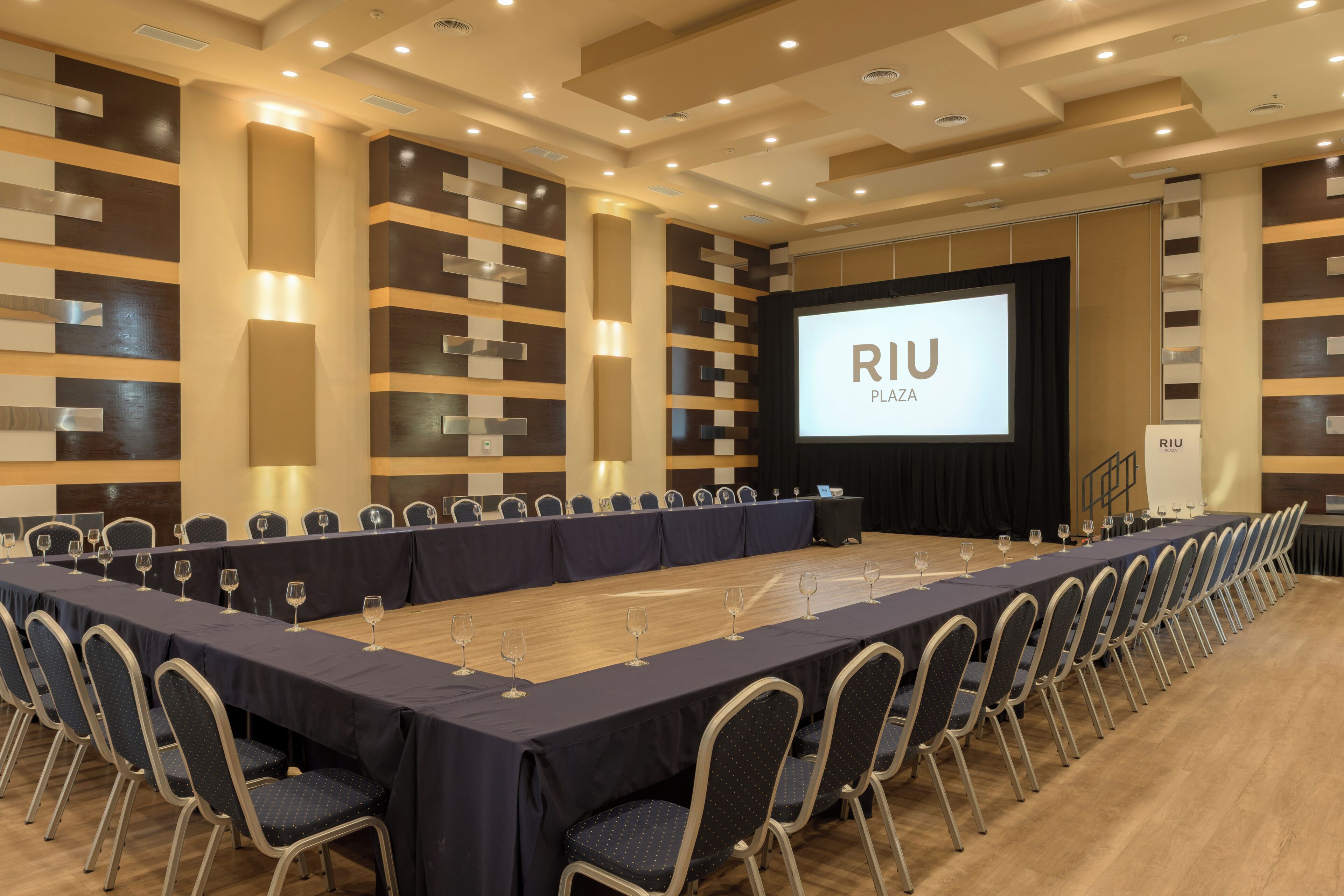 A conference room with a long table, chairs, and a screen displaying 'RIU PLAZA'.