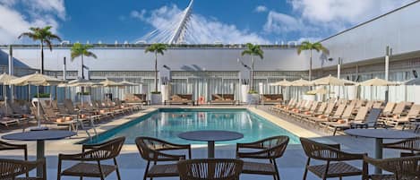 Zona junto a la piscina con sillones, mesas y sombrillas, con vistas a un puente moderno.