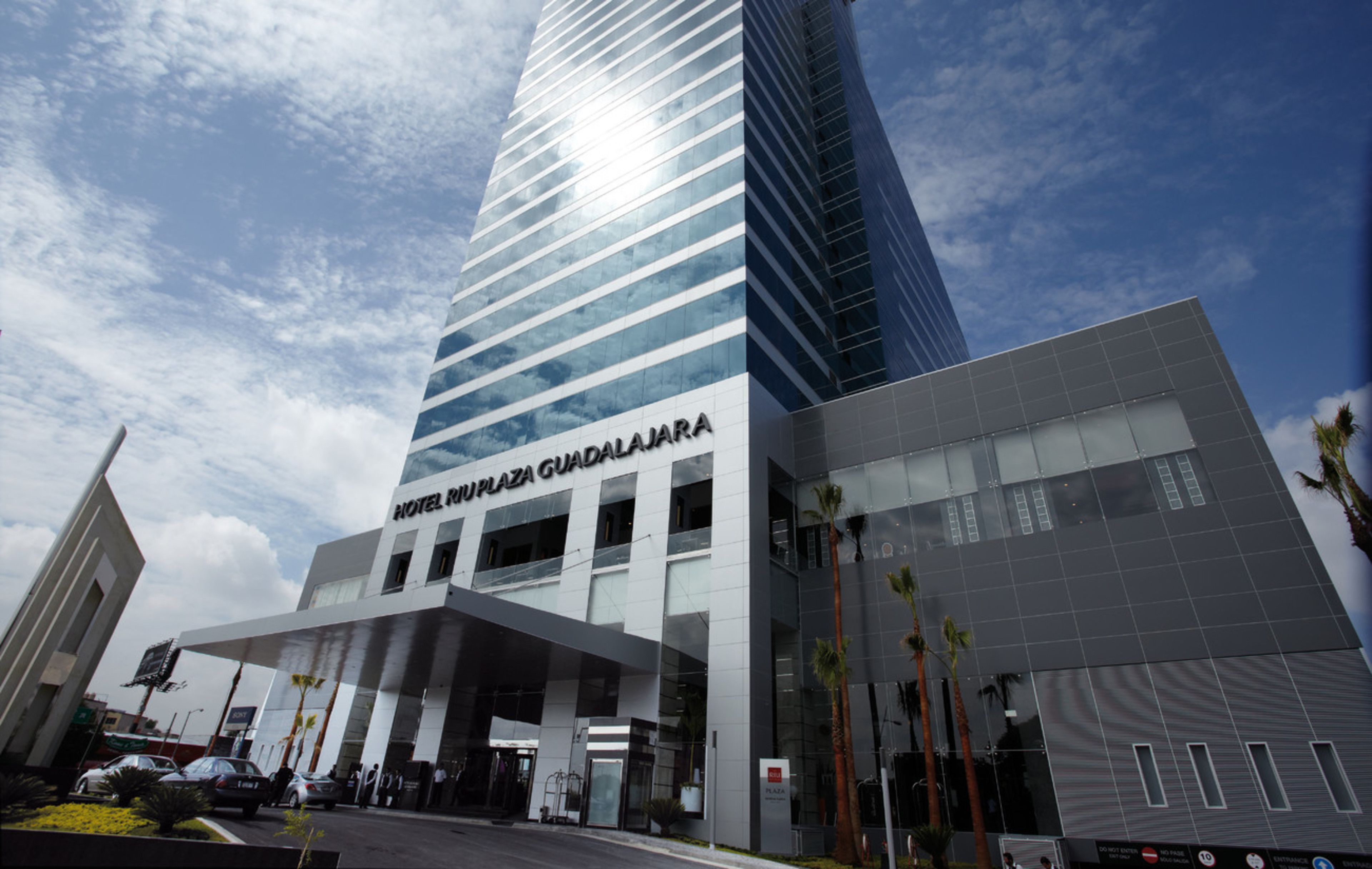 A modern hotel building with a prominent name sign, a covered entrance, and a parking area.