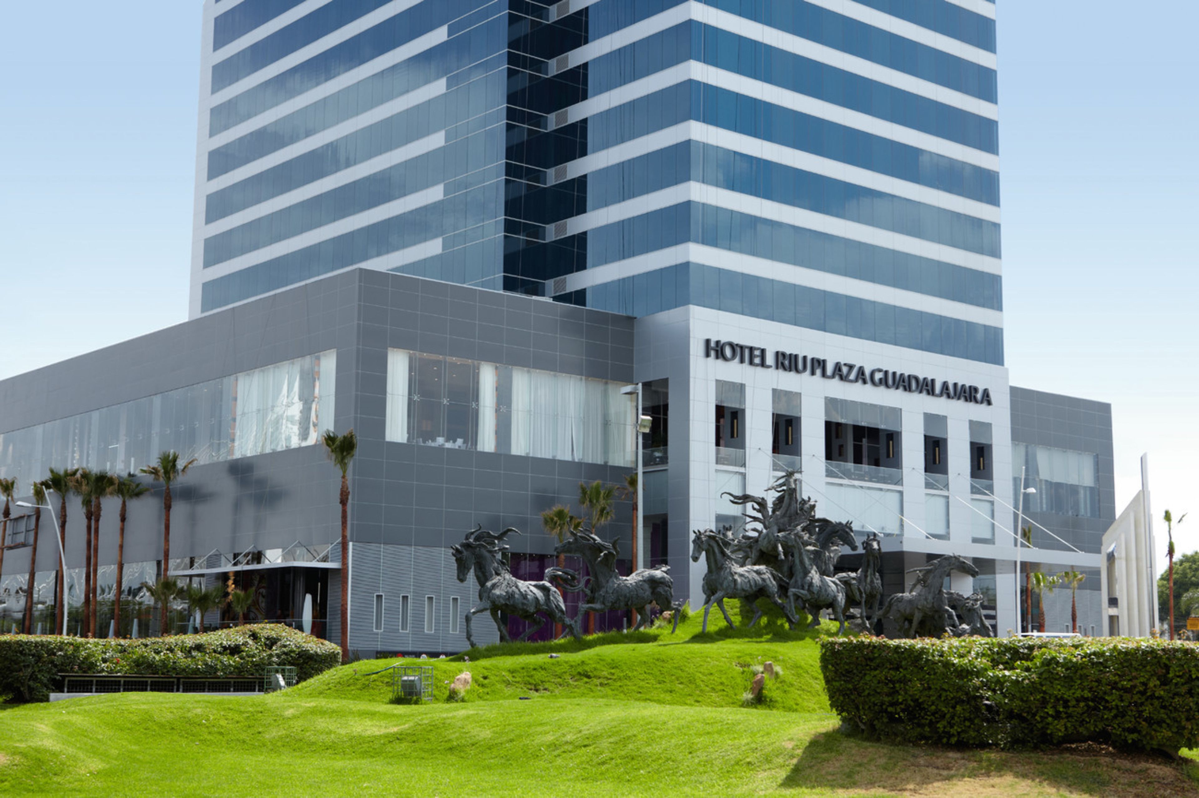 Un hotel moderno con una amplia fachada de vidrio, una entrada destacada y una escultura de caballos al frente.