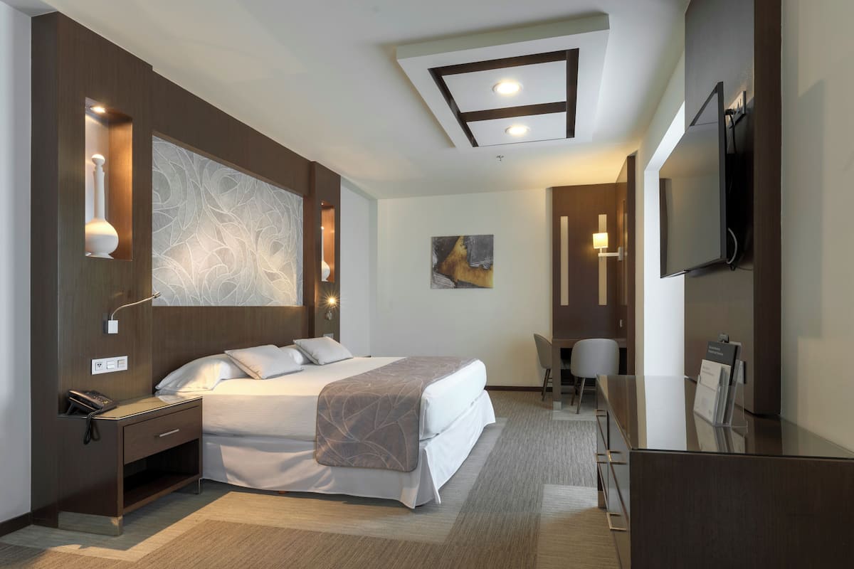 Une chambre d’hôtel moderne dotée d’un grand lit, d’une table de chevet, d’un bureau, d’une chaise et d’un téléviseur à écran plat fixé au mur.