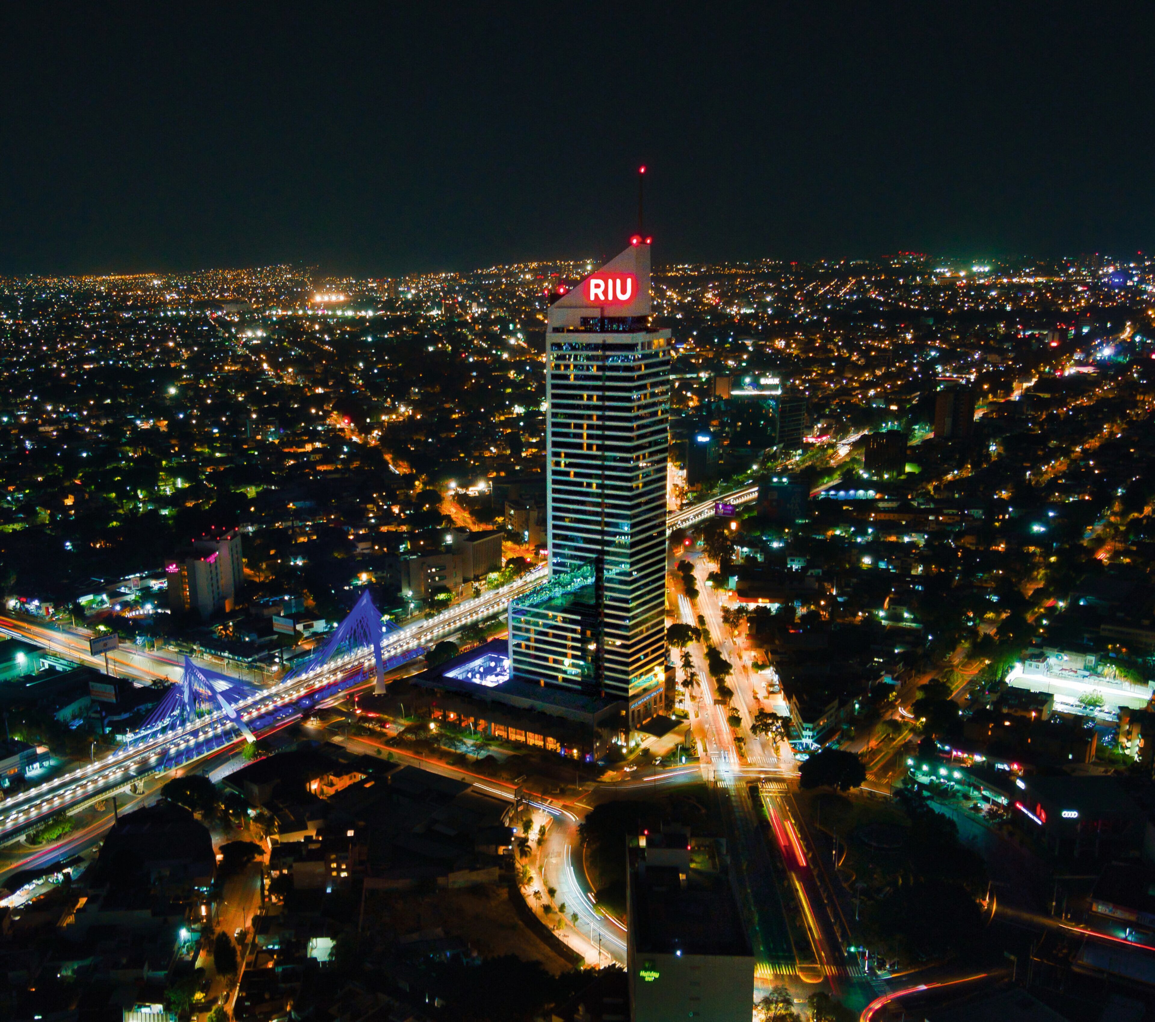 Un hotel de gran altura con un llamativo letrero 'RIO' en la parte superior, rodeado de una ciudad densamente poblada de noche.