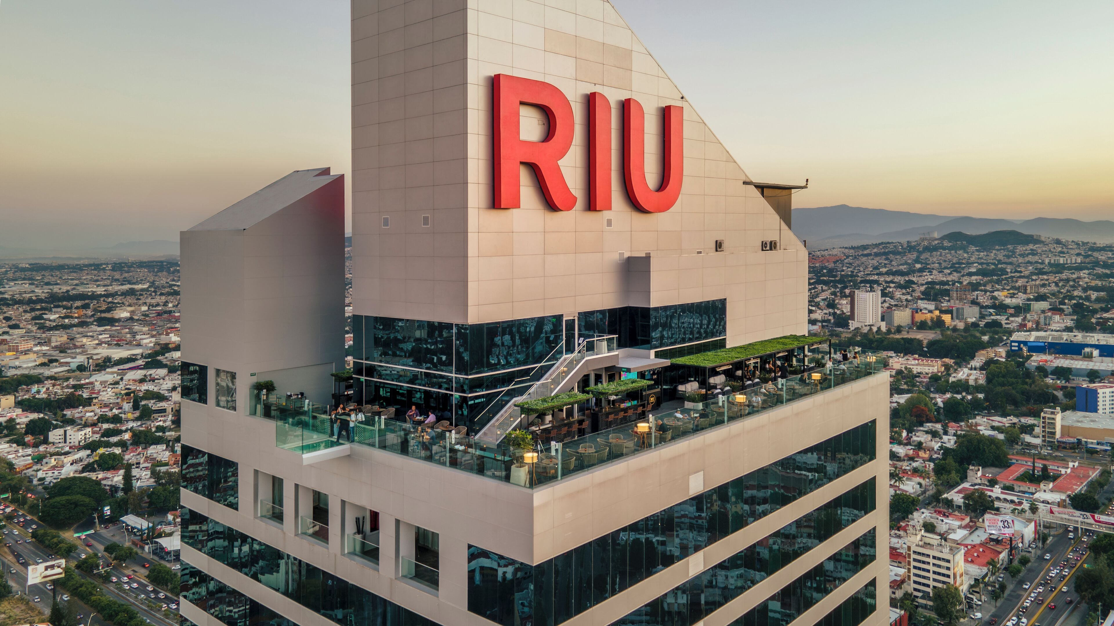 A modern building with a prominent 'RIU' sign, featuring a rooftop bar with outdoor seating and a glass facade.