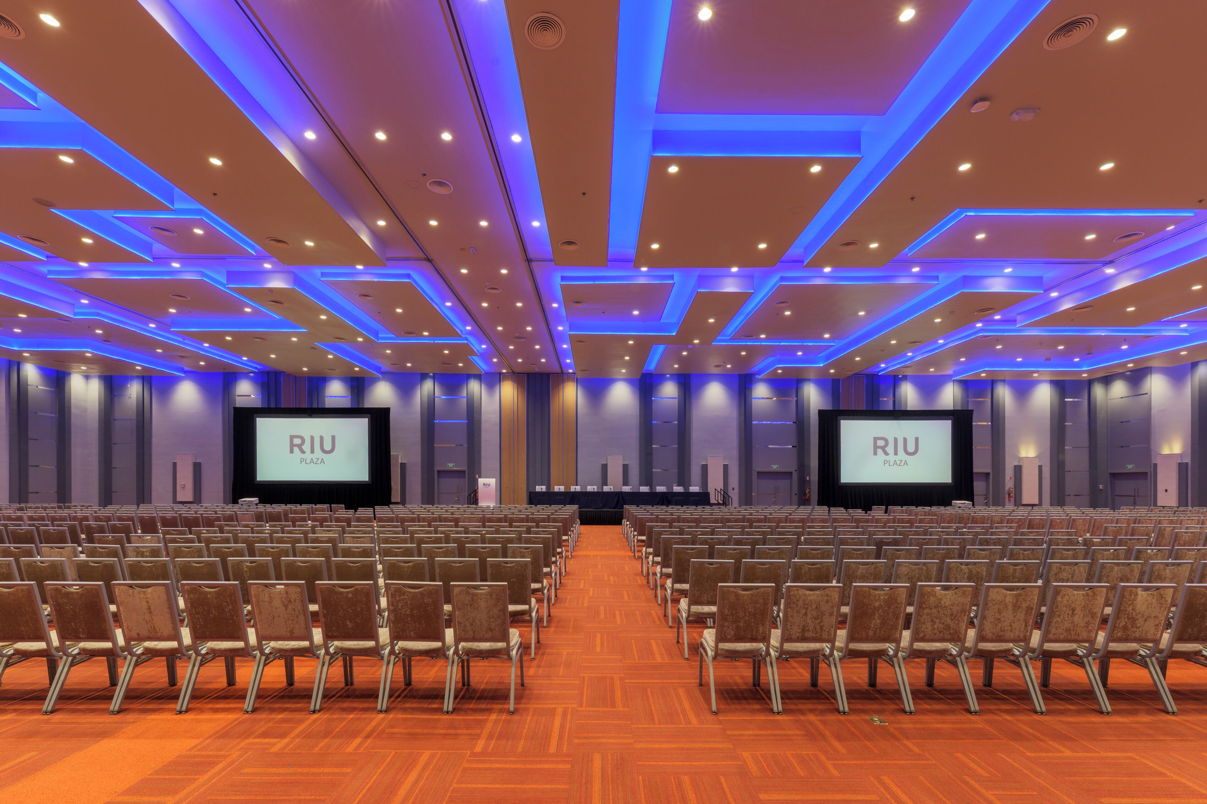 A conference room with rows of chairs, two large screens, and a wooden floor.