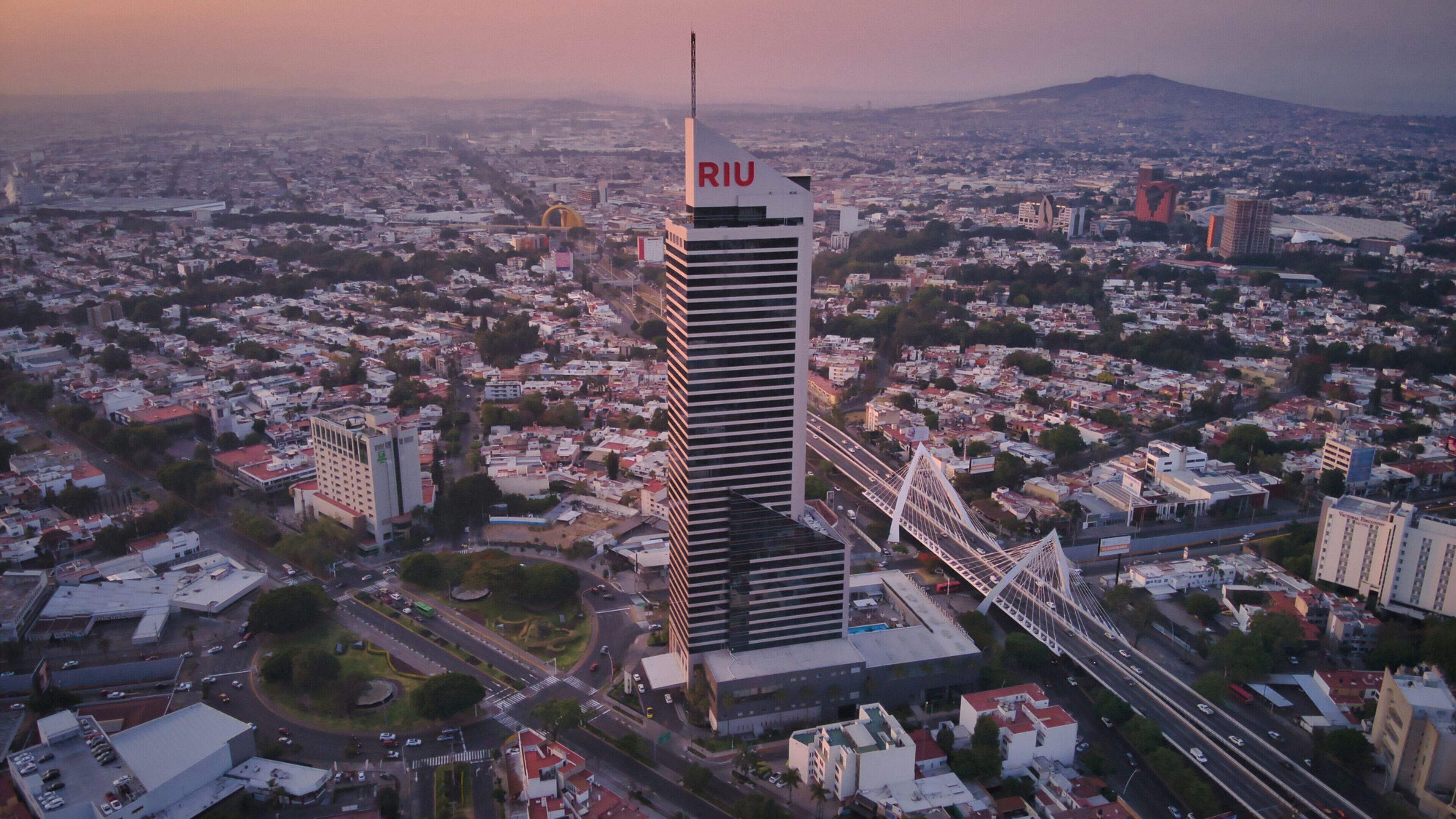 A high-rise building with the acronym "RIU" on its facade, surrounded by a densely populated urban area with various buildings and a bridge in the foreground.