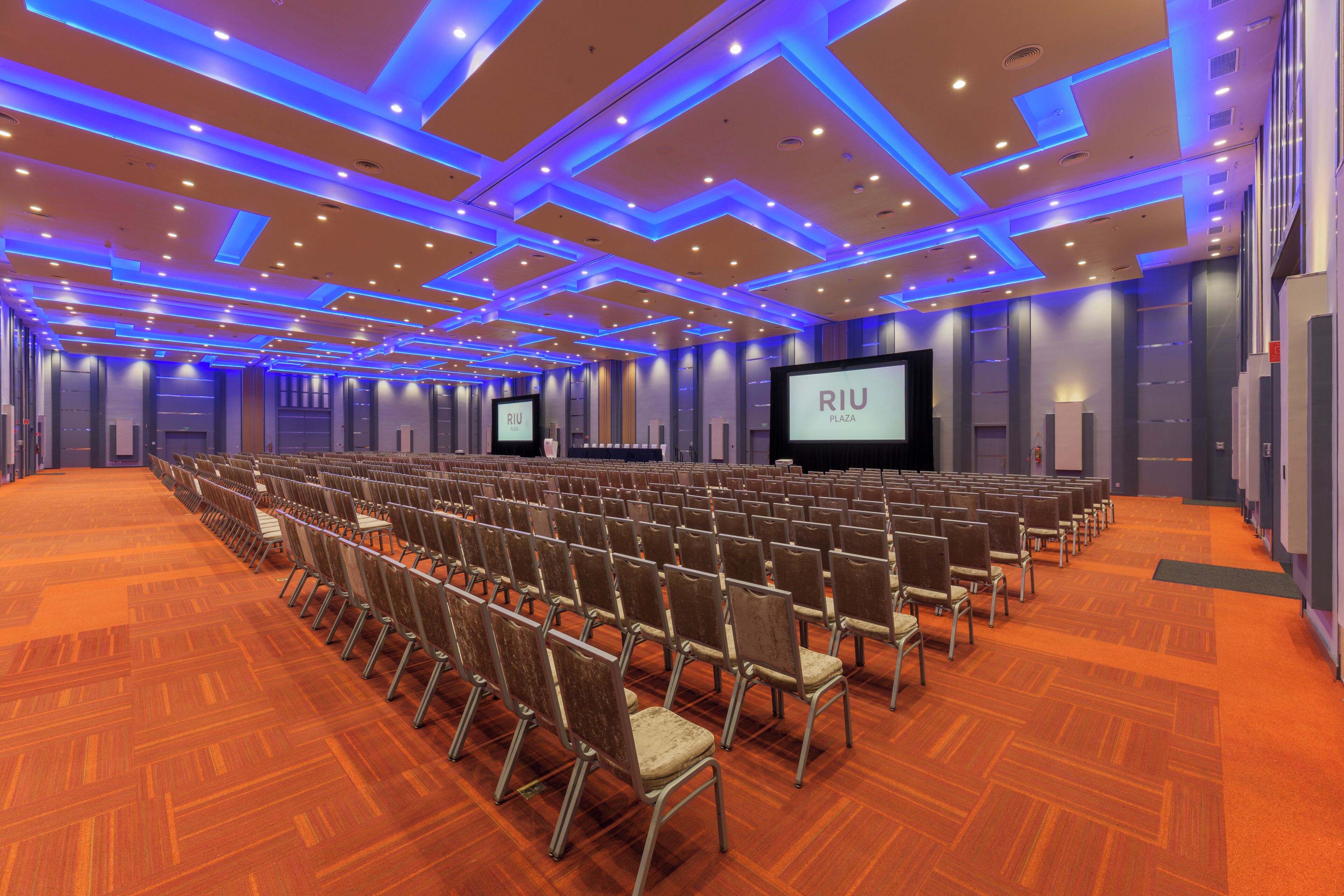 A conference room with rows of chairs, a large screen, and a 'RIU Plaza' sign.