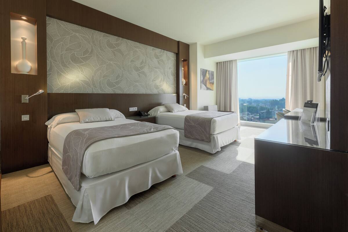 Une chambre d’hôtel avec deux lits, une grande fenêtre donnant sur la ville, un minibar et un téléviseur à écran plat.