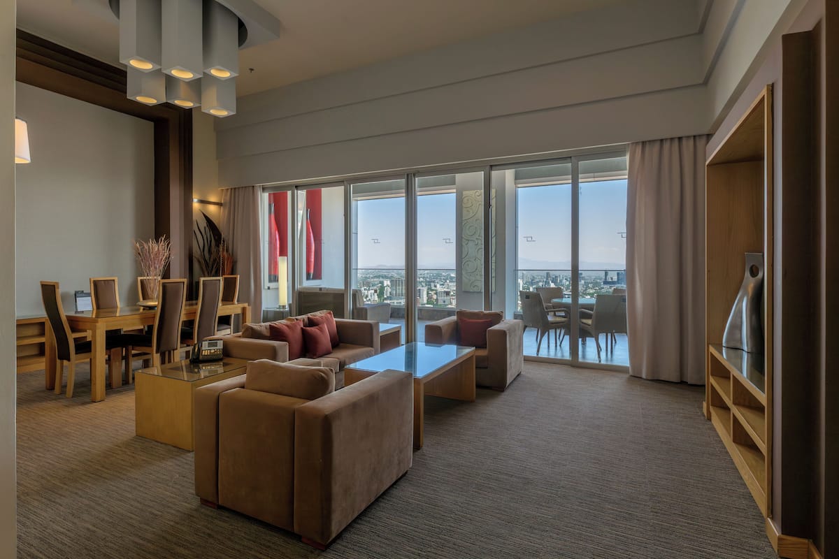 Une chambre d’hôtel moderne dotée d’une grande fenêtre, d’un coin salon avec des canapés et une table basse, d’une table à manger avec des chaises, et d’un téléviseur placé à droite.