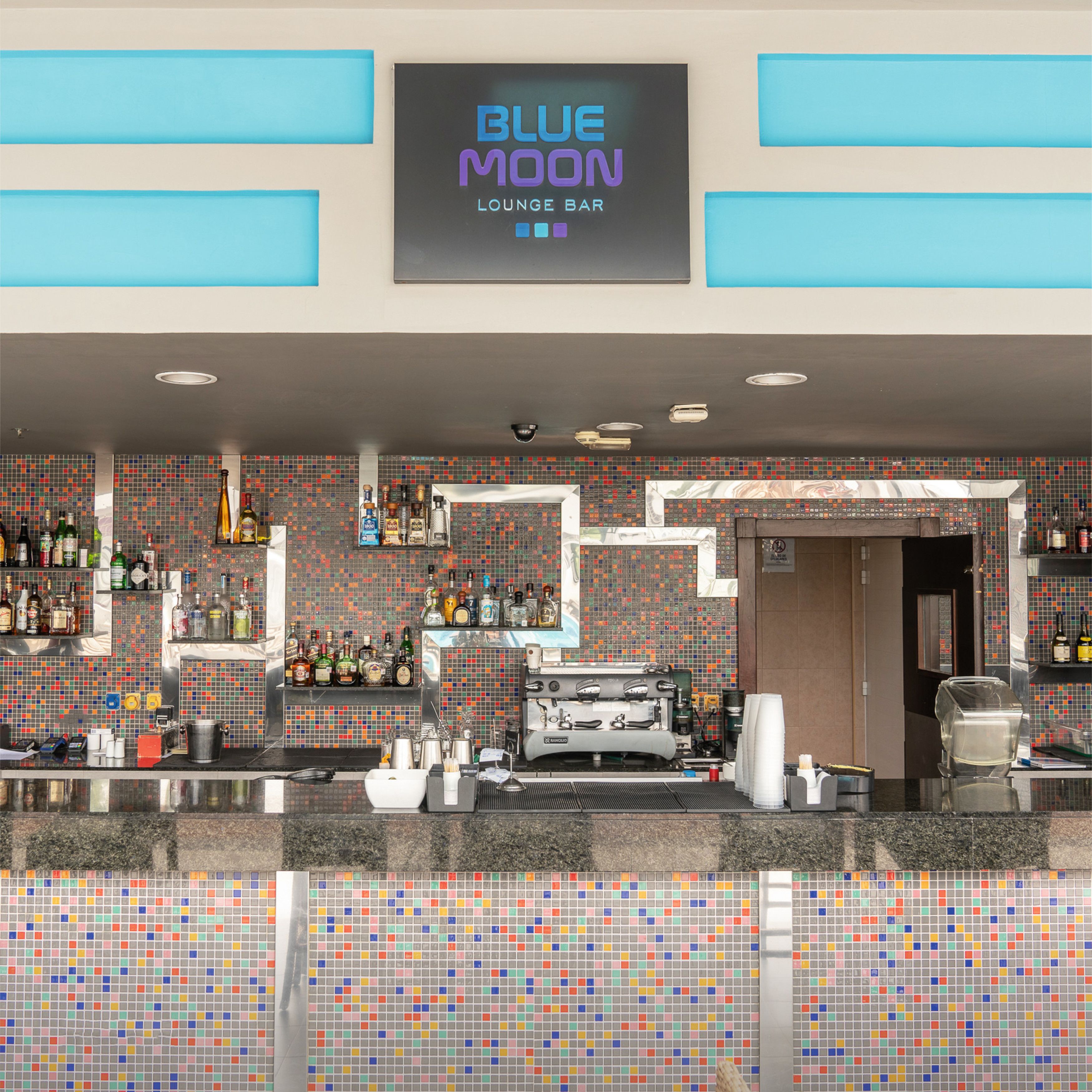 A bar area with a tiled wall, shelves with bottles, and a coffee machine.