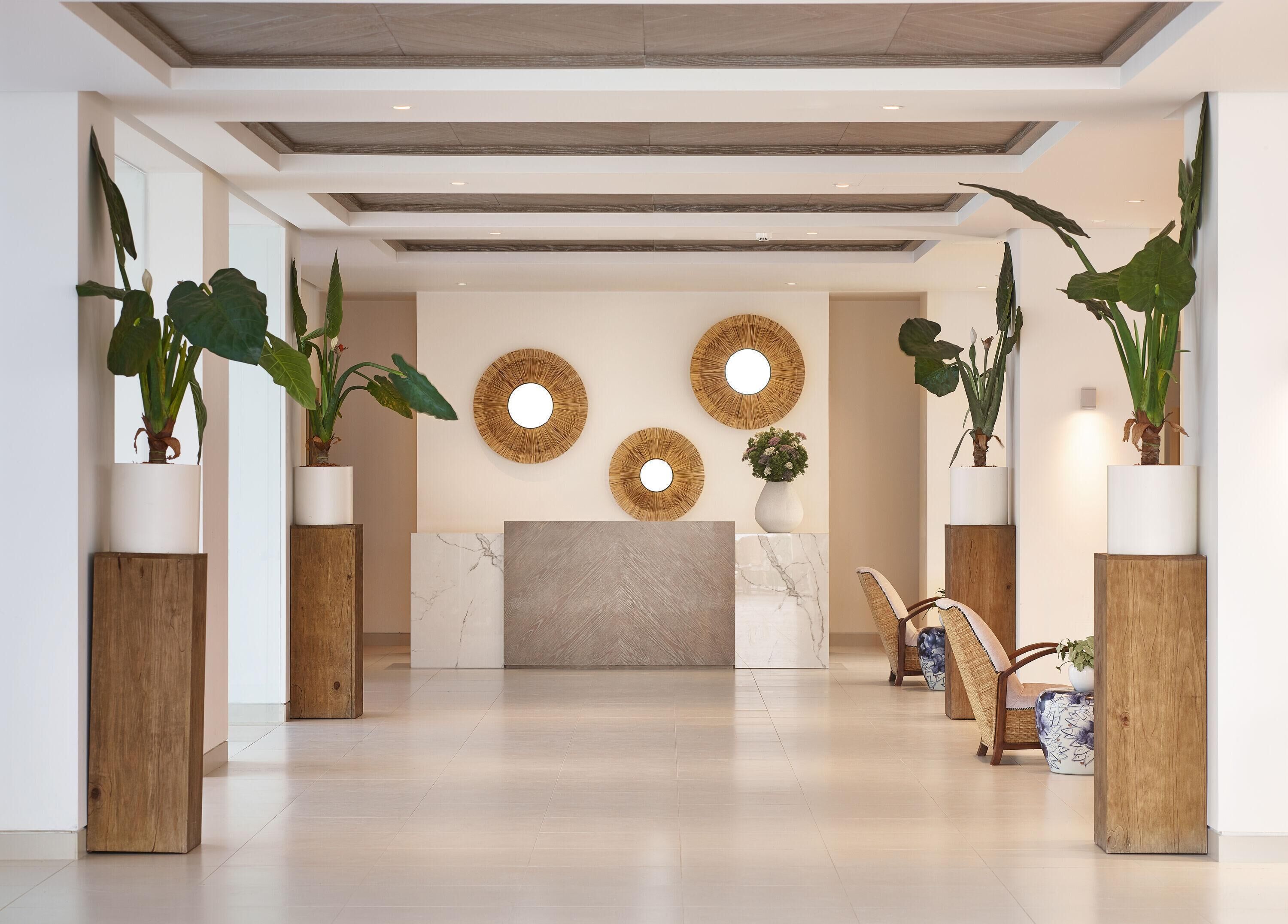 Un espace d’accueil moderne avec un comptoir de réception en marbre, des piliers en bois, de grandes plantes en pot et des miroirs décoratifs fixés aux murs.