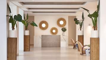 Un espace d’accueil moderne avec un comptoir de réception en marbre, des piliers en bois, de grandes plantes en pot et des miroirs décoratifs fixés aux murs.