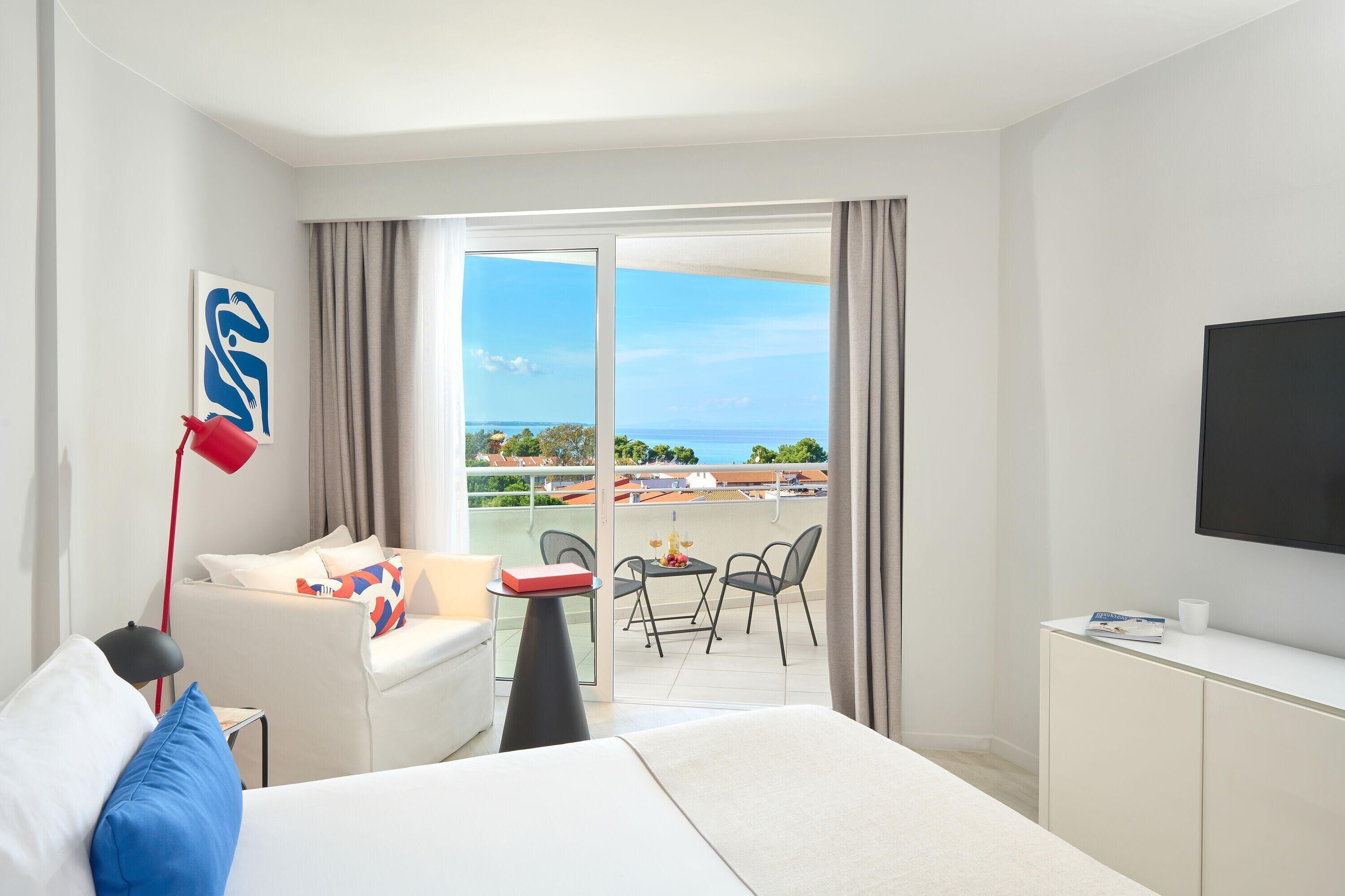 A modern hotel room with a balcony, a flat-screen TV, a sofa, a coffee table, and a desk.