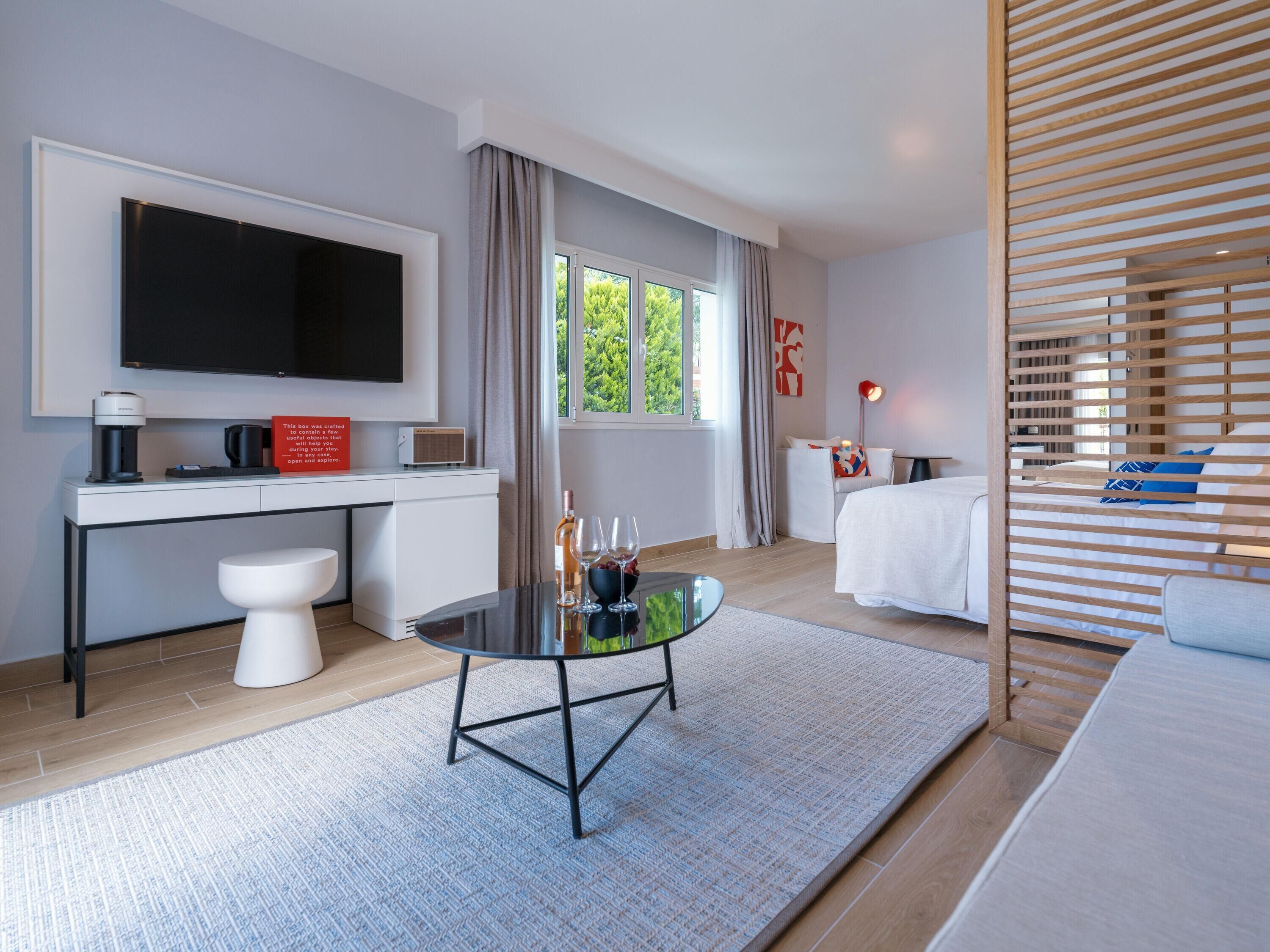 A modern hotel room with a flat-screen TV, a desk with a coffee maker, a bedside table with wine glasses, a bed with wooden slats, and a large window with curtains.