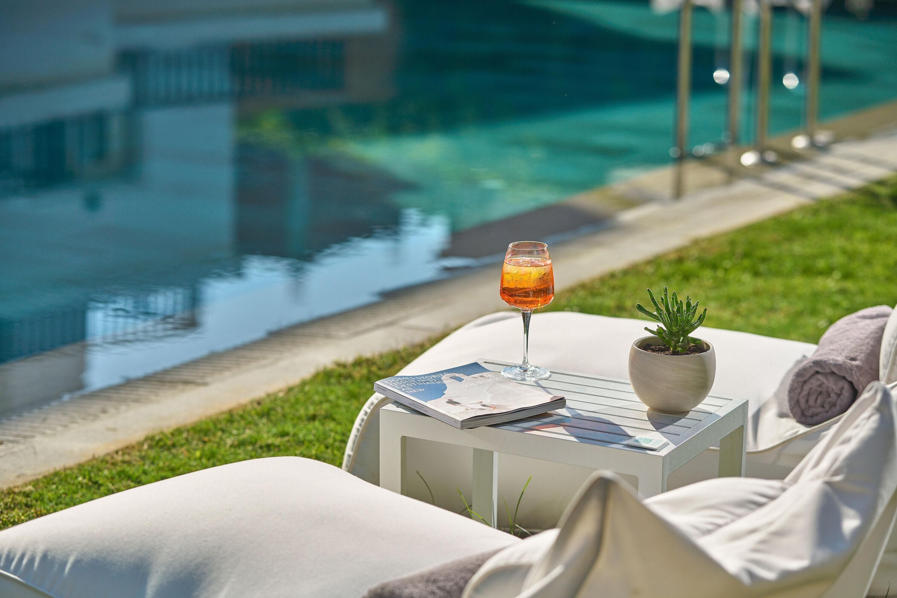 Zona de descanso junto a la piscina con una bebida de naranja, una revista y una planta en maceta sobre una mesita.