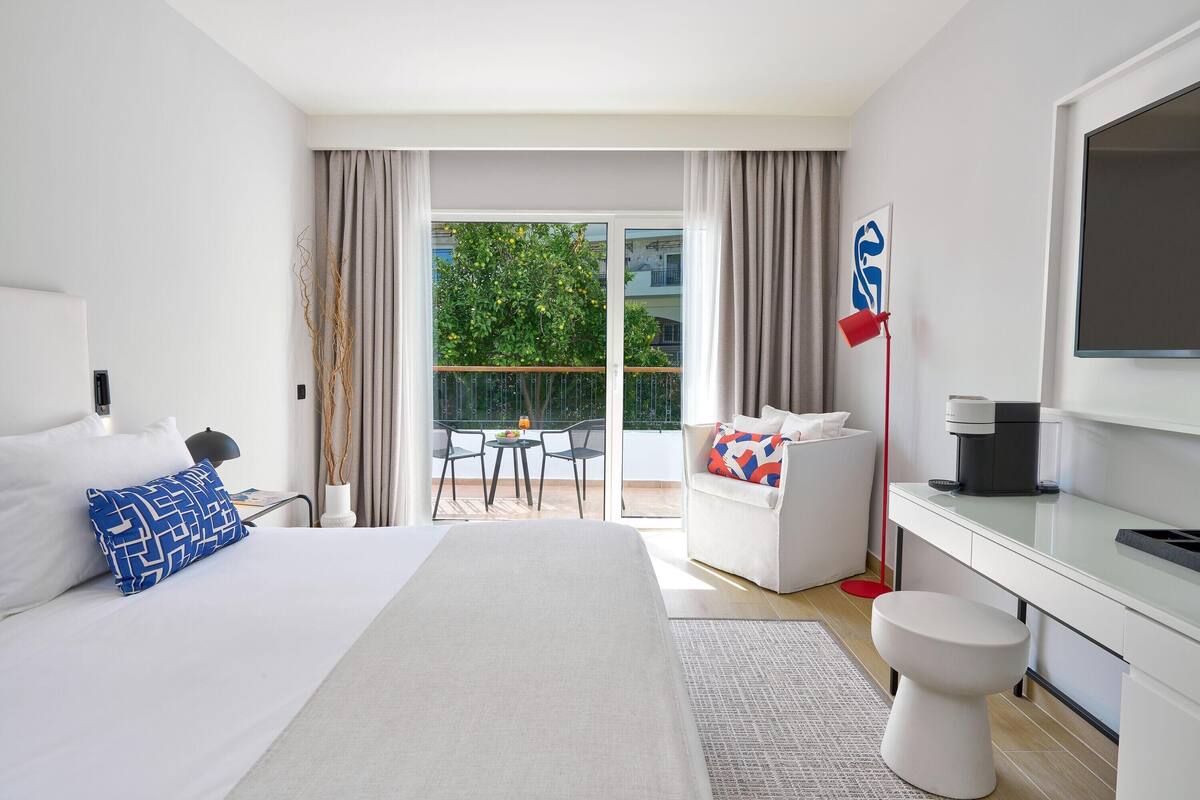 Ein modernes Hotelzimmer mit einem großen Bett, einem Schreibtisch, einem Stuhl, einer Toilette und einem Balkon mit Tisch und Stühlen.