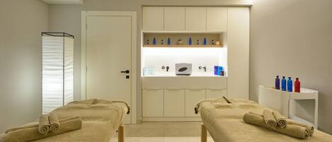 Un cuarto de spa con dos camillas de masaje, un armario blanco con botellas y un espejo.