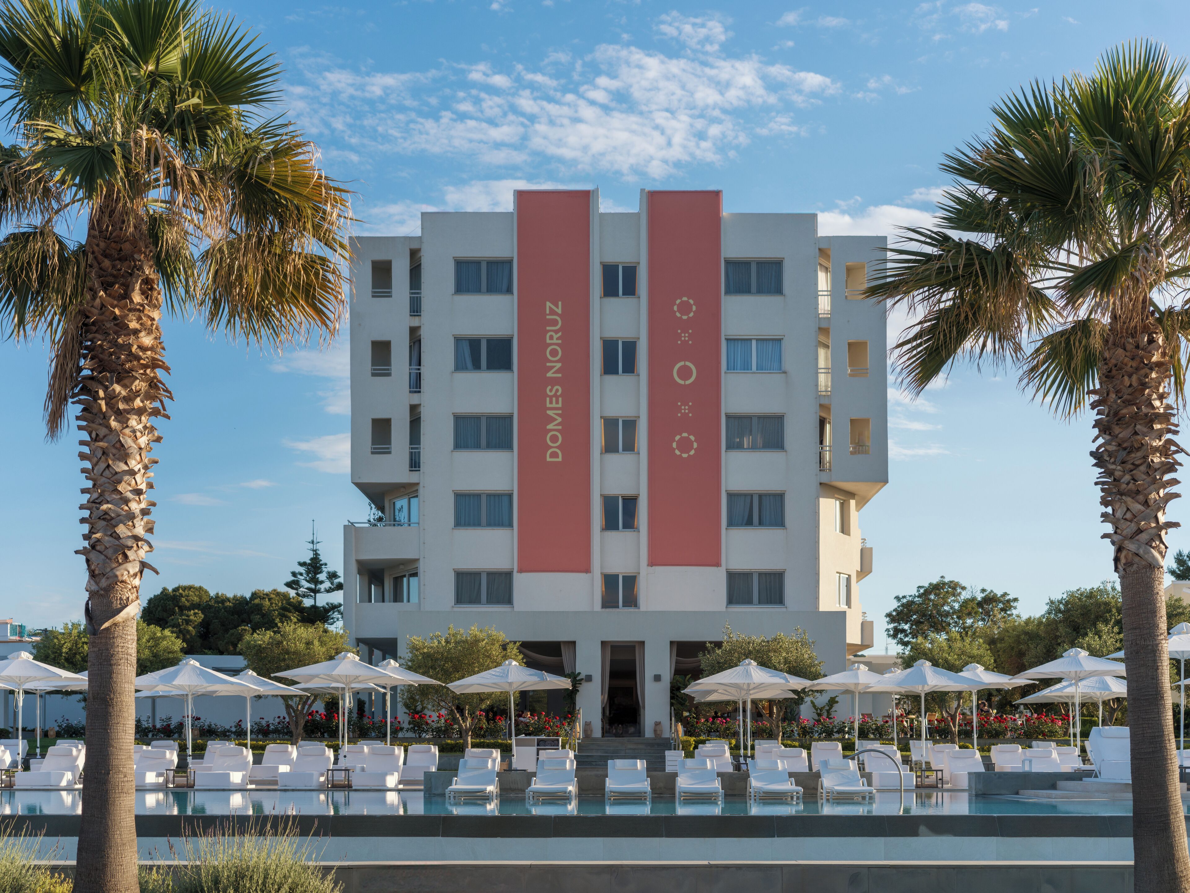 A hotel with a pool area, palm trees, and white umbrellas.