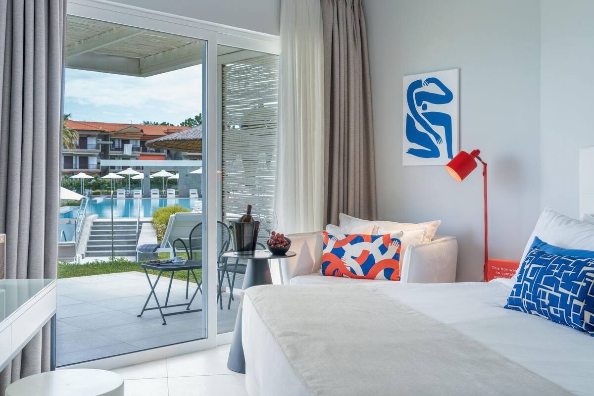 Ein modernes Hotelzimmer mit Balkon, einem Bett mit gemusterten Kissen, einer roten Stehlampe und Blick auf einen Pool und Gebäude.