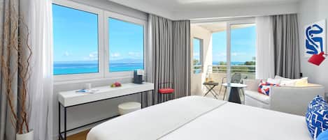 A hotel room with a large bed, a desk, a chair, and a balcony with a view of the sea.