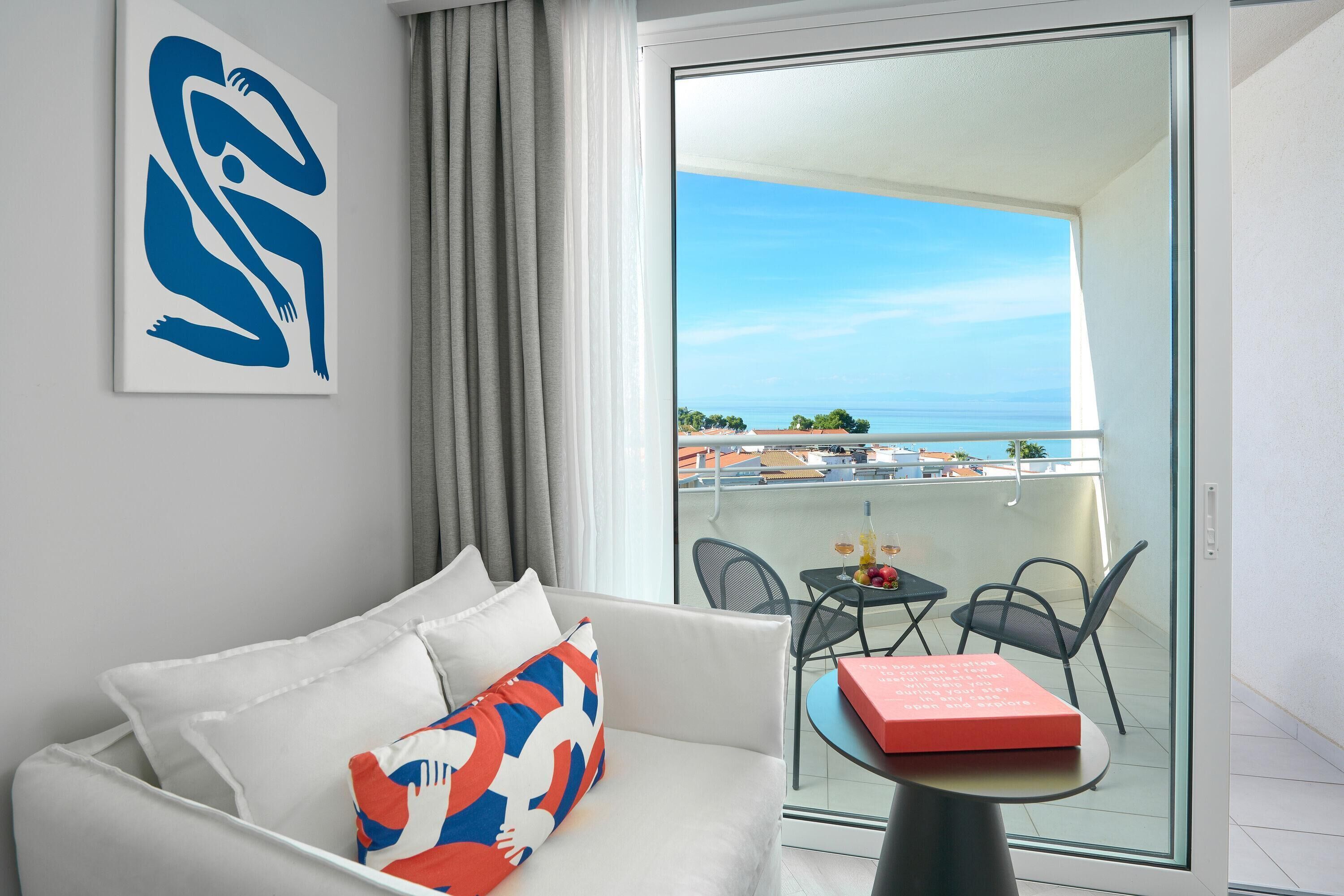 A modern hotel room with a white sofa, a colorful pillow, a glass table with a red book, and a balcony with a view of the sea.