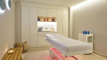 A modern room with a massage table, a small wooden table, a basket, and a shelf with bottles.
