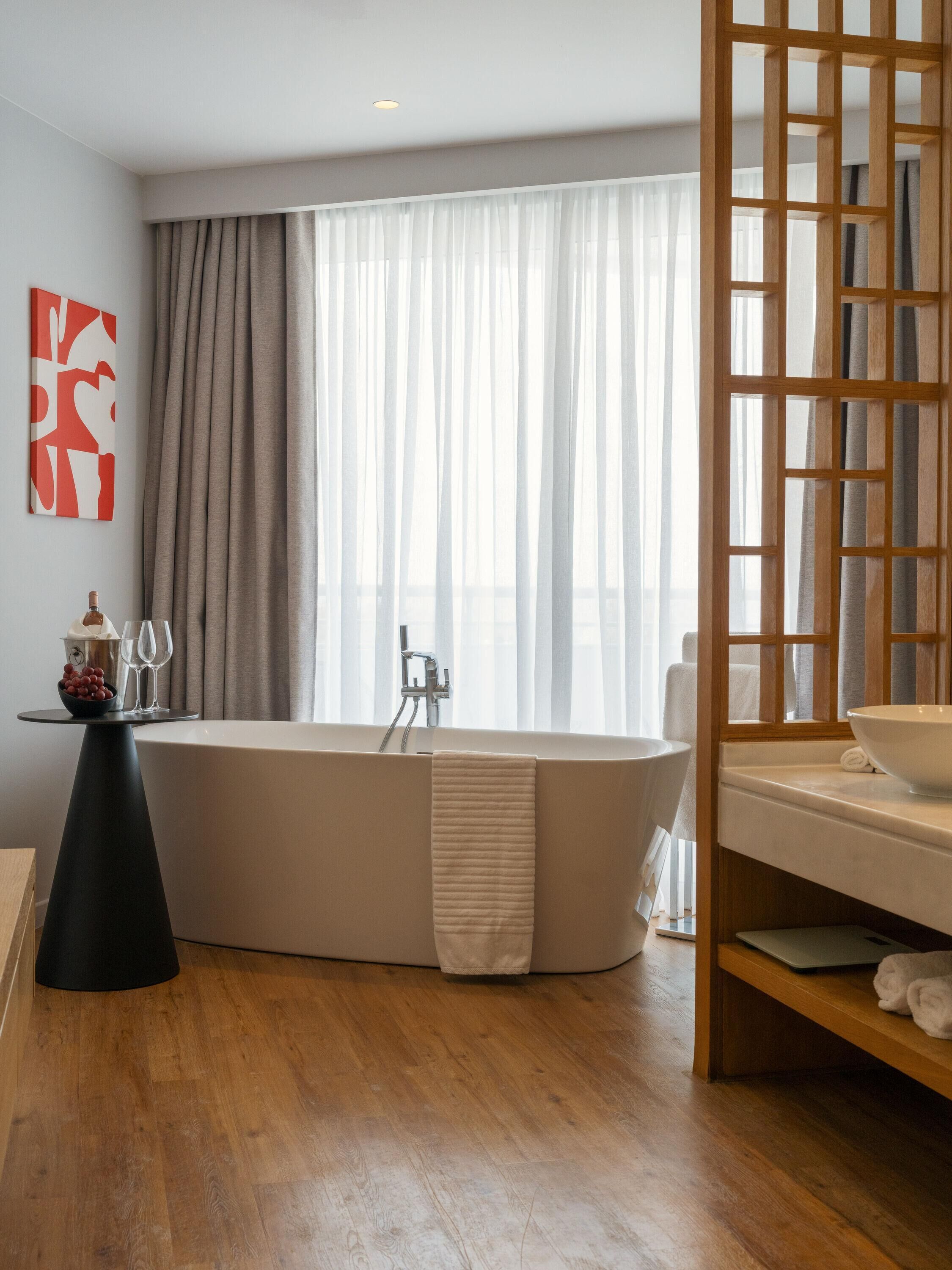 A modern bathroom with a freestanding bathtub, a wooden floor, and a large window with sheer curtains.