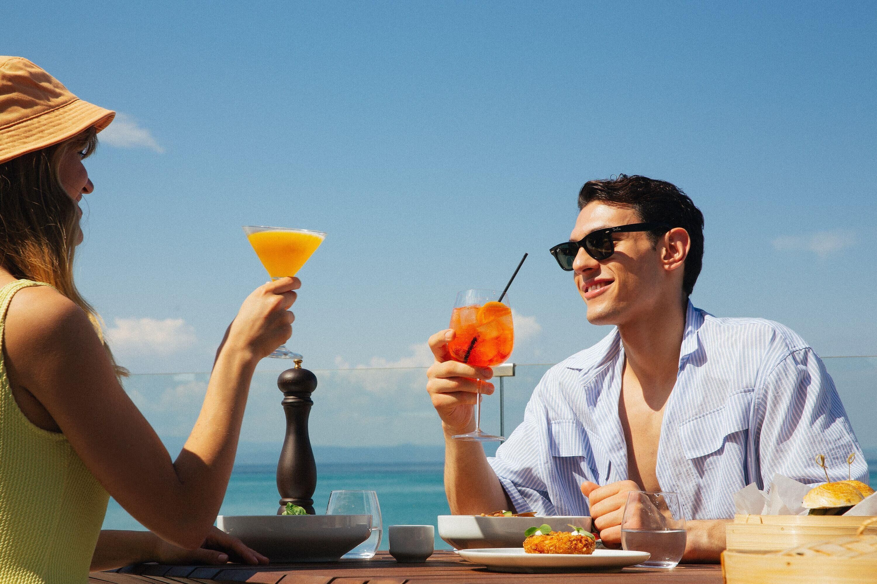 Un couple est assis à une table avec vue sur l’océan, savourant un cocktail et un repas.