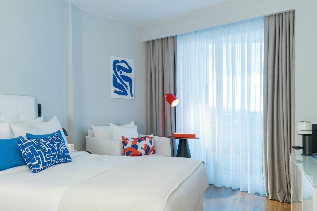Ein modernes Hotelzimmer mit einem großen Bett, einem Nachttisch mit Lampe und Blick auf die Stadt durch leichte Vorhänge.