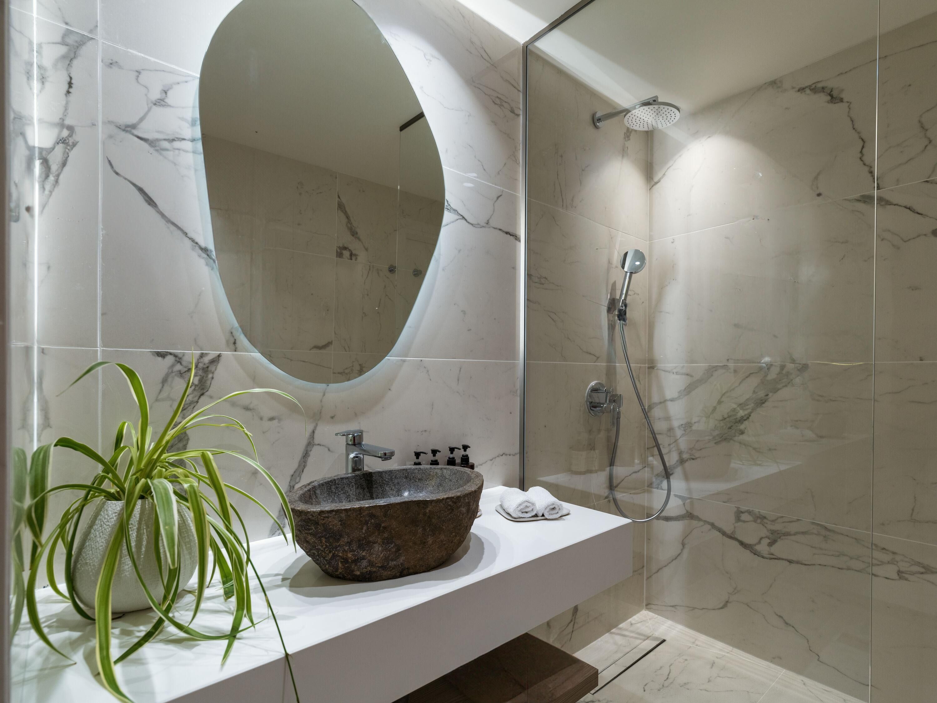 A bathroom with a marble-tiled wall, a round mirror, a stone sink, a shower, and a potted plant.