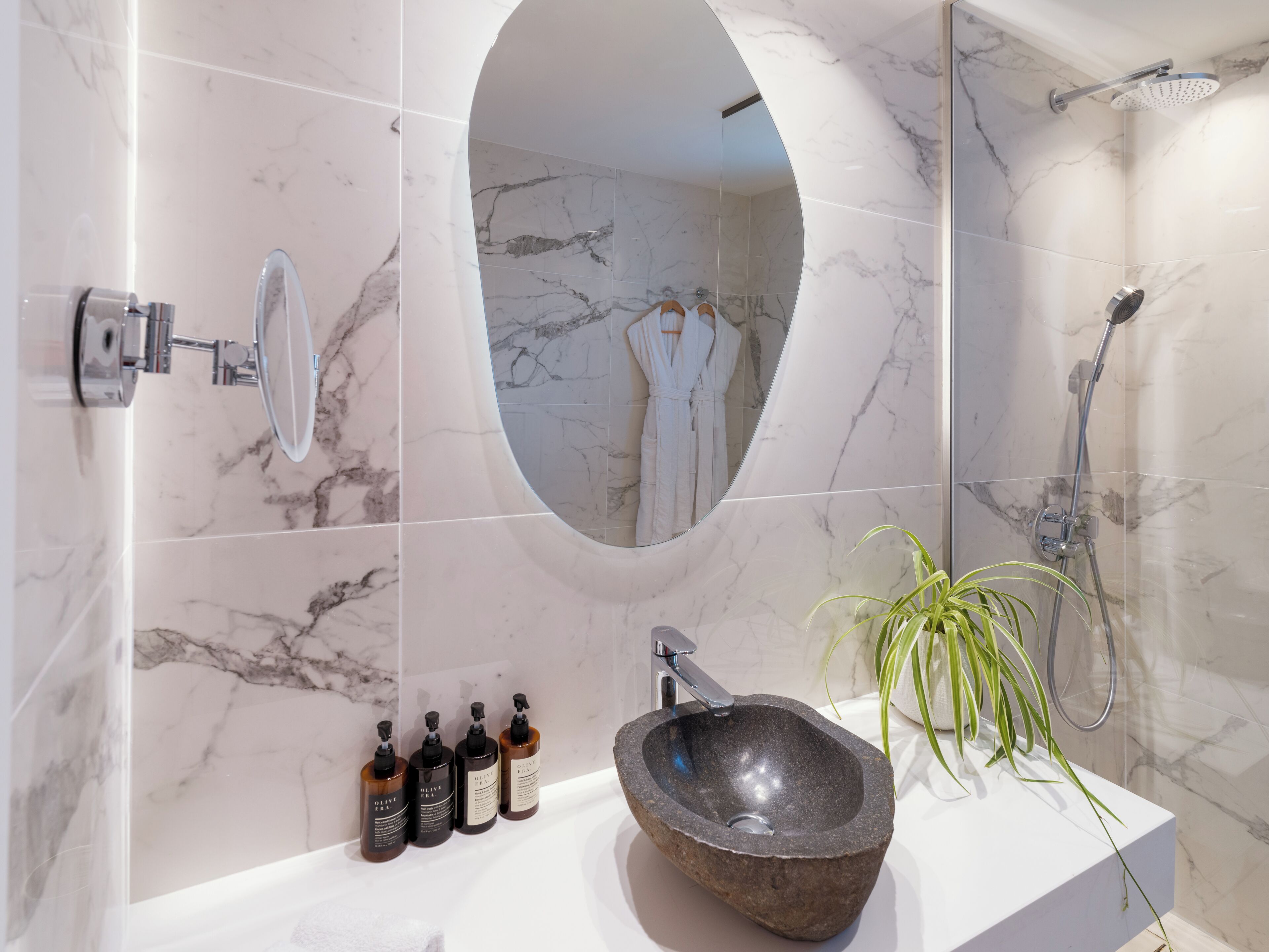A modern bathroom with a marble backsplash, a large oval mirror, a stone sink, and a shower area.