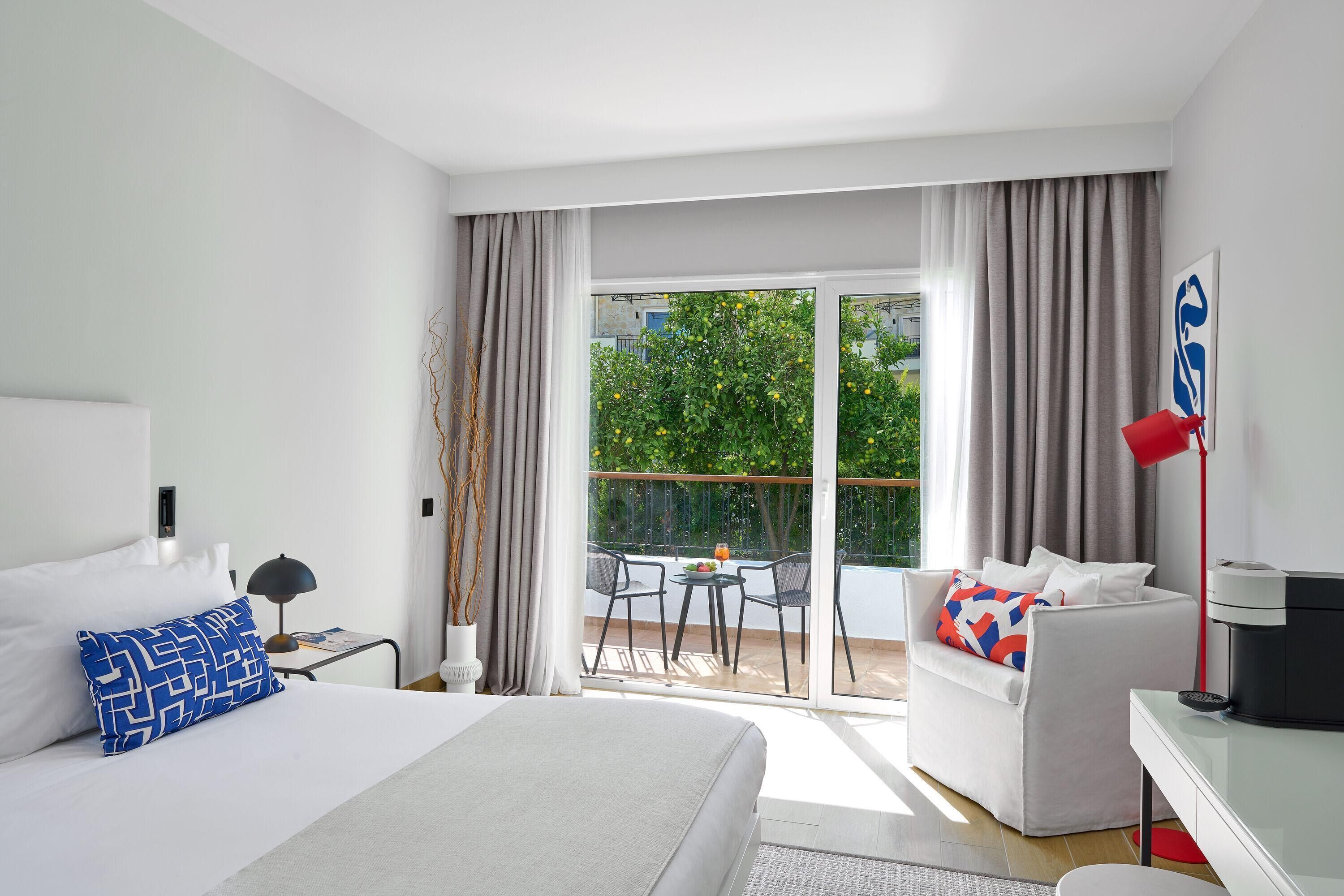 A modern hotel room with a bed, a sofa, a desk, and a balcony with a table and chairs.