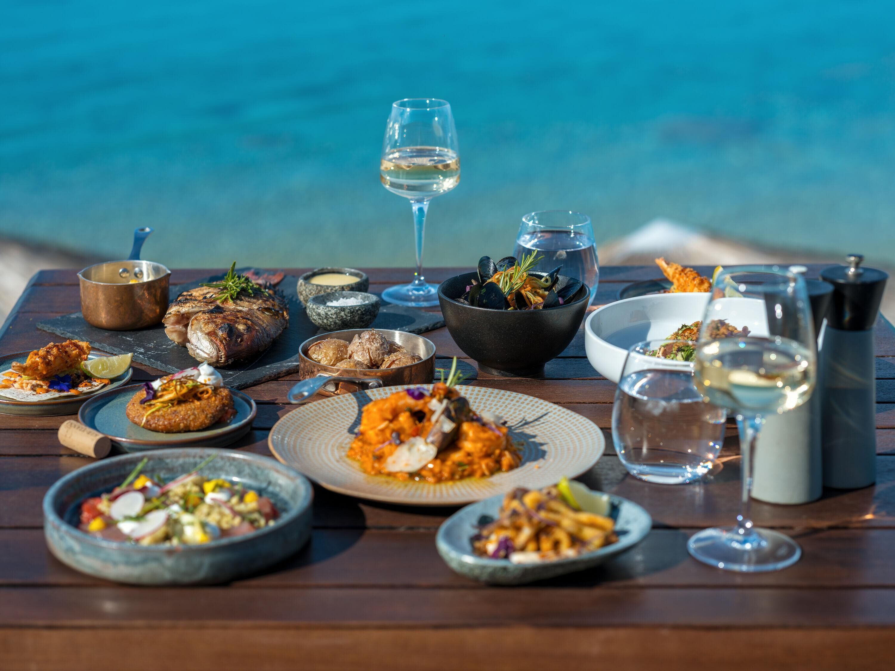 A table set with various dishes, a glass of white wine, and a view of the sea.