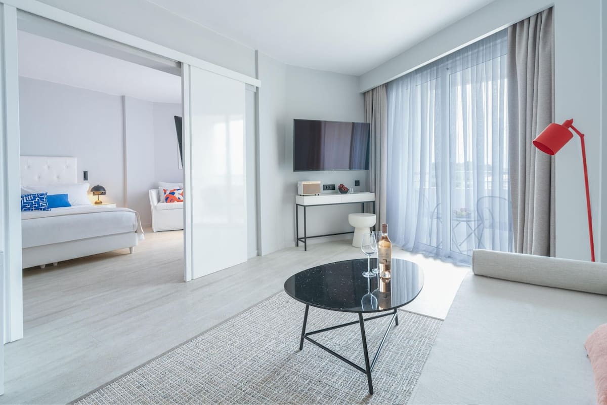 Ein modernes Hotelzimmer mit Bett, Sofa, Esstisch, Fernseher und einem Fenster mit Vorhängen.