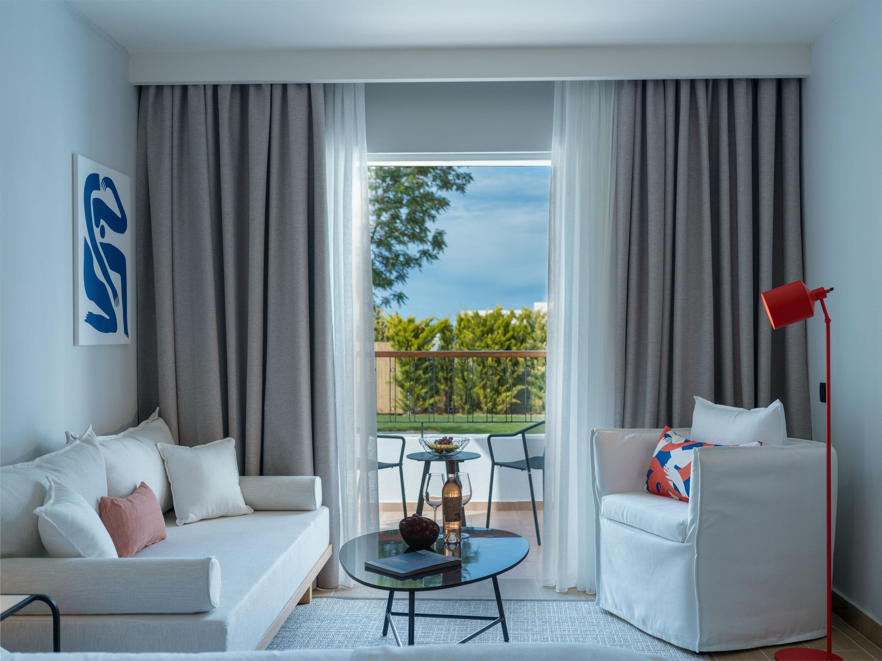 A modern hotel room with a balcony, a sofa, a chair, a small table with a bottle and glasses, and a red floor lamp.