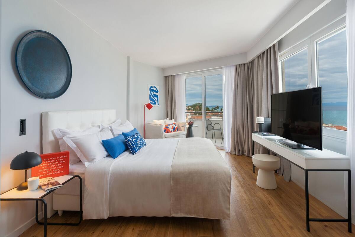 Ein modernes Hotelzimmer mit einem großen Bett, einem Schreibtisch, einem Fernseher und einem Balkon mit Meerblick.