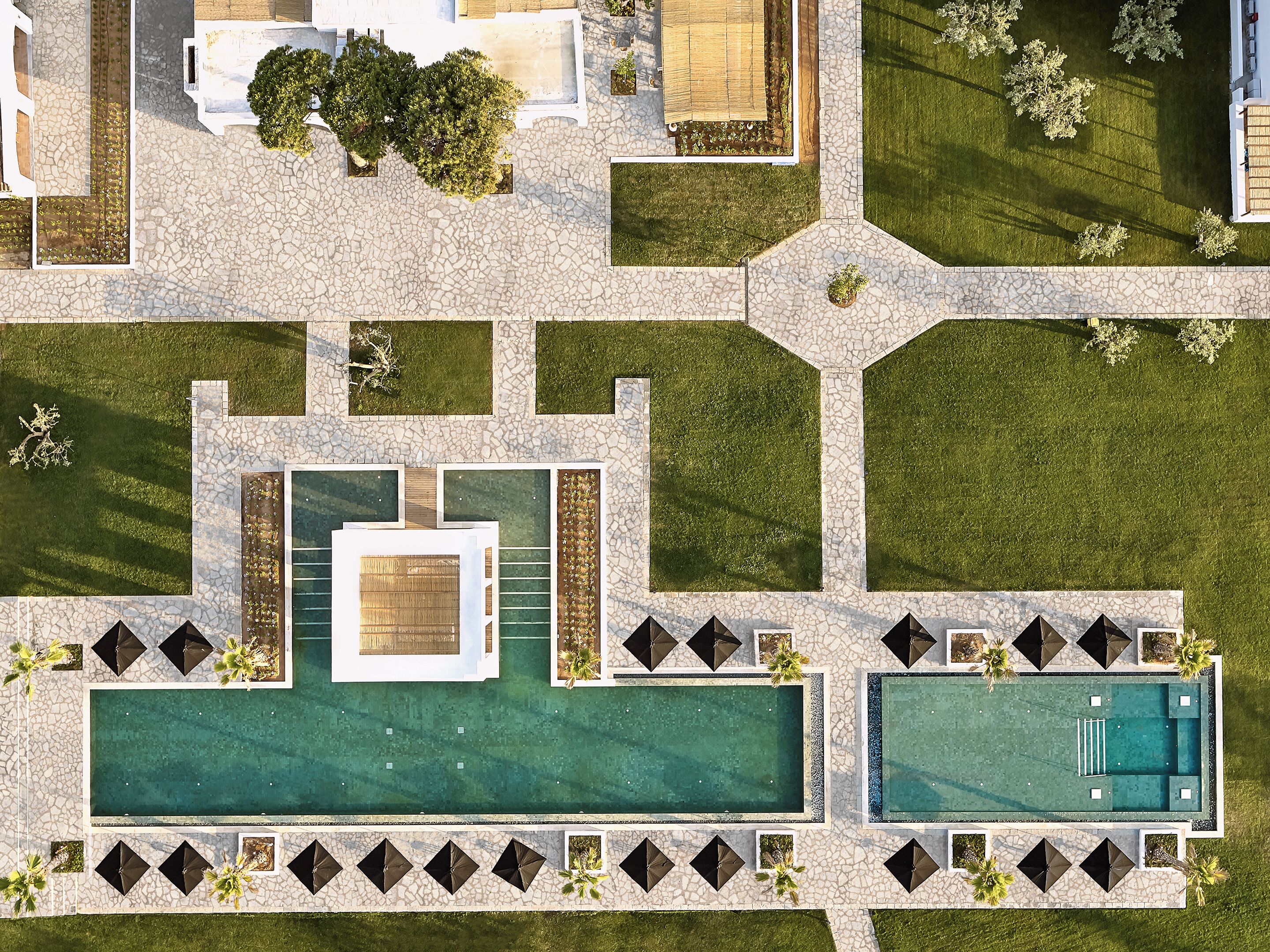 Vista aerea di un resort con piscina centrale, diverse piscine più piccole e una zona verdeggiante con vialetti e vegetazione.