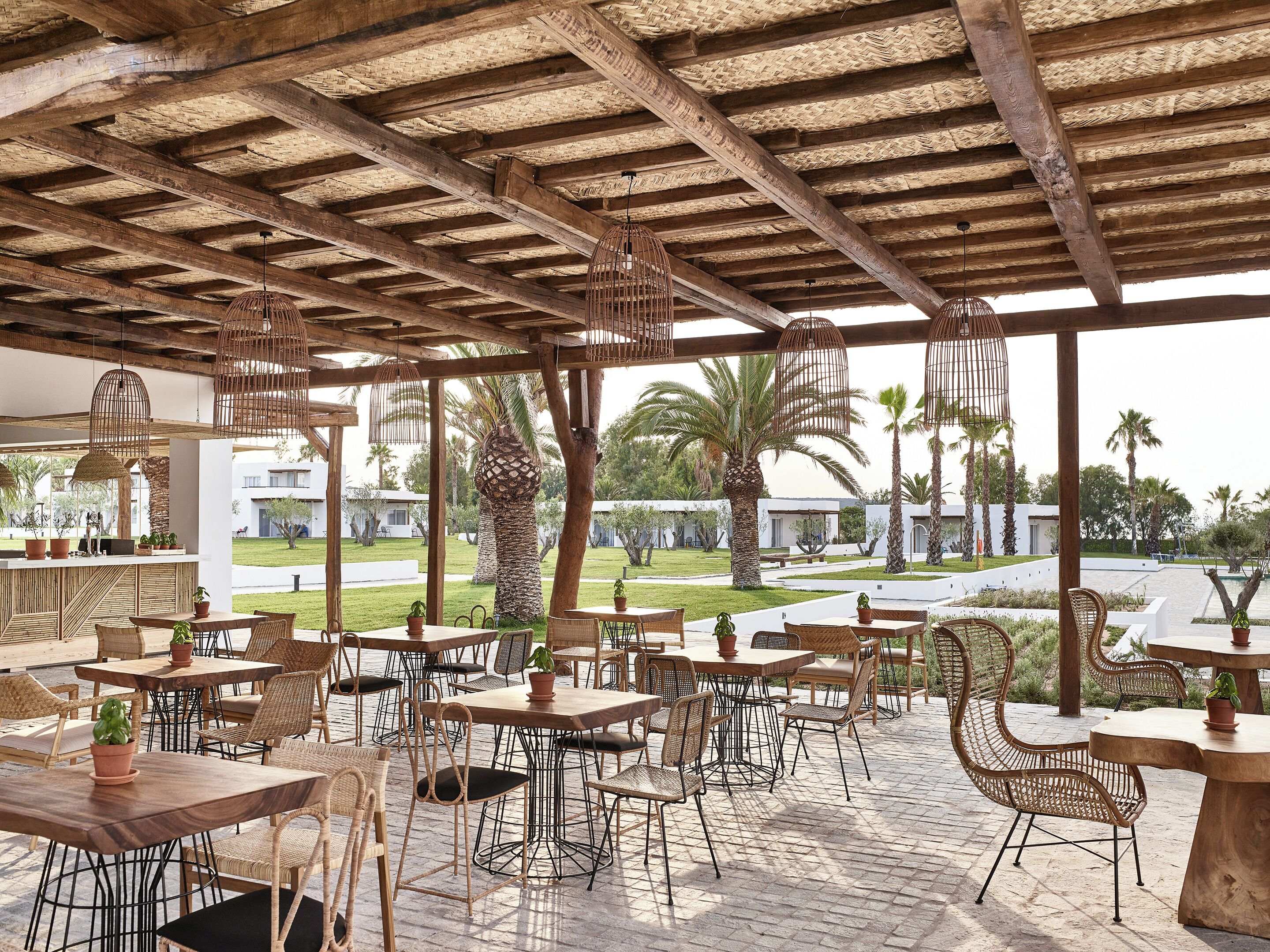 An outdoor patio with wooden tables and wicker chairs, a stone floor, and a view of a residential area with palm trees.