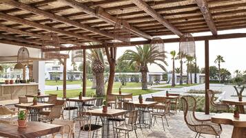 An outdoor patio with wooden tables and wicker chairs, a stone floor, and a view of a residential area with palm trees.