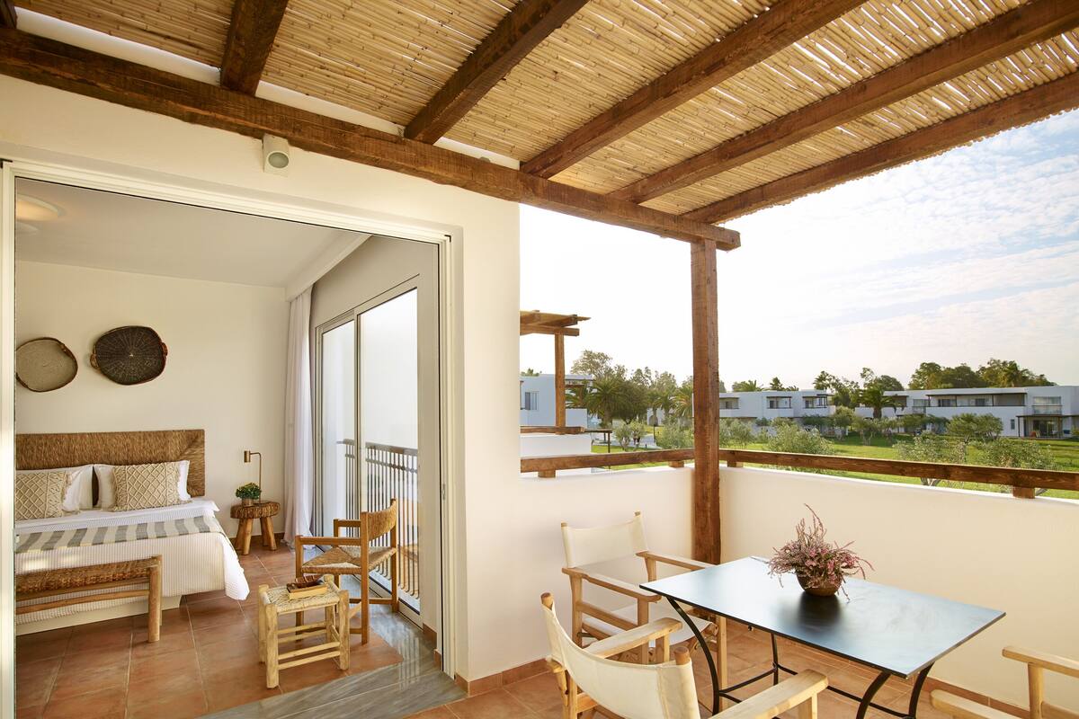 A balcony with a wooden table and chairs, a bed with a wicker headboard, and a view of a residential area.