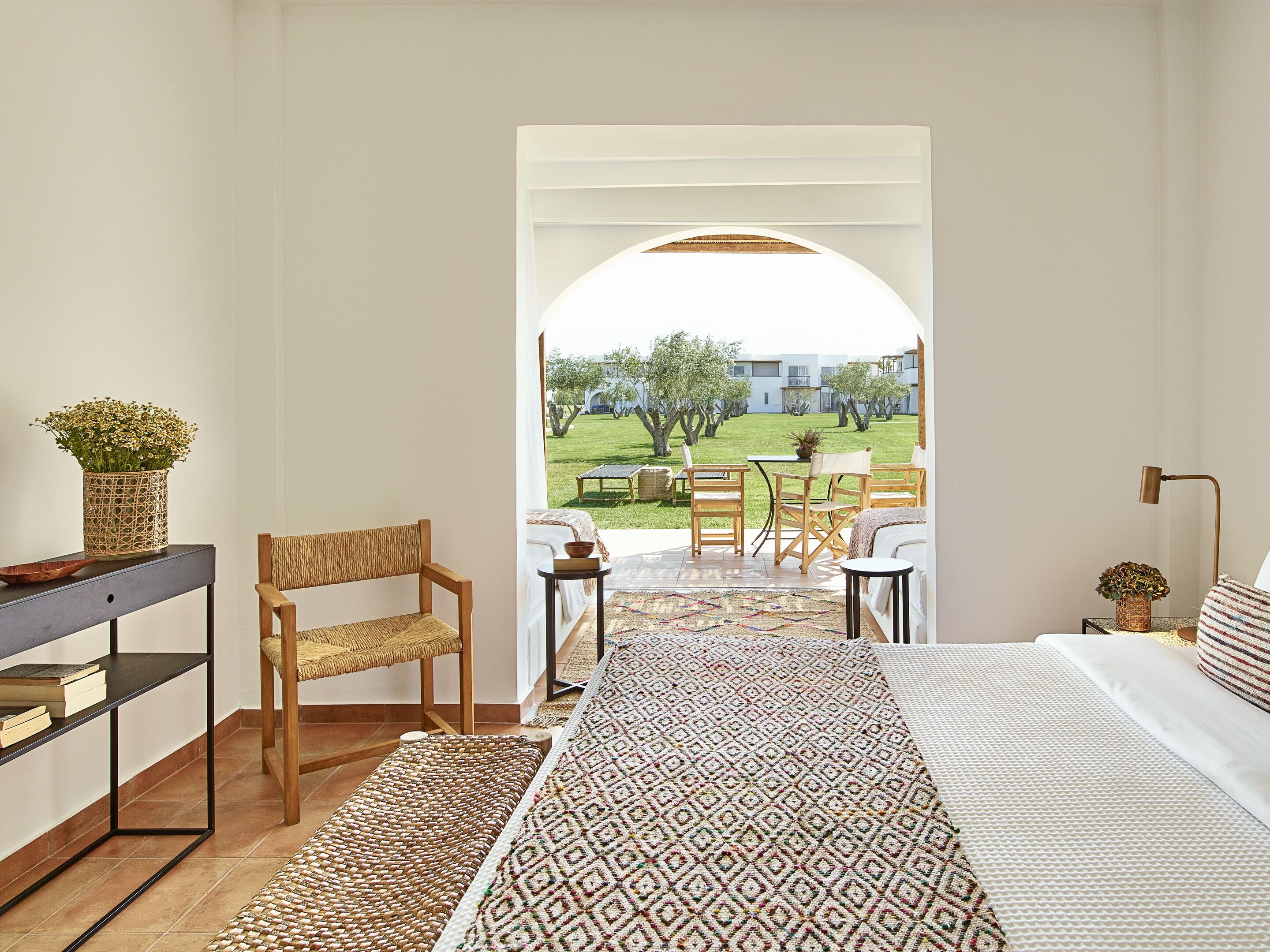 A room with a bed, a wooden chair, a small table, and a view of a garden and buildings outside.