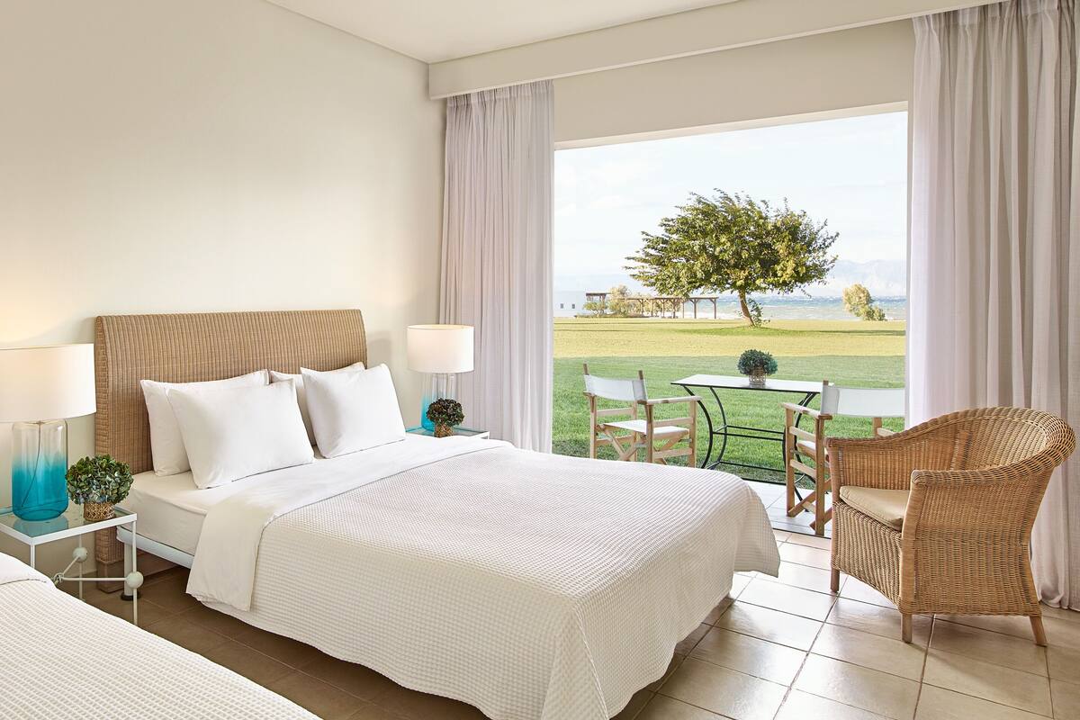 A hotel room with a bed, a nightstand, a table with chairs, and a view of a grassy field and a tree.