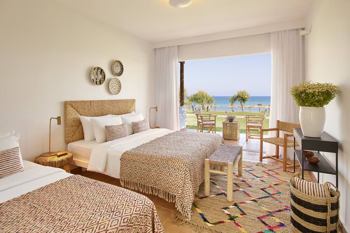 A hotel room with two beds, a large window with a view of the beach, and a balcony with outdoor seating.